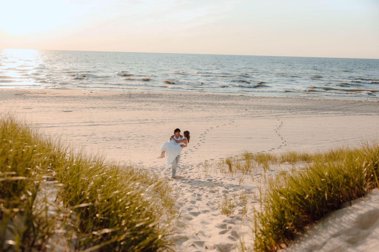 Couple embracing on a sandy beach at sunset, surrounded by dune grass and gentle ocean waves.