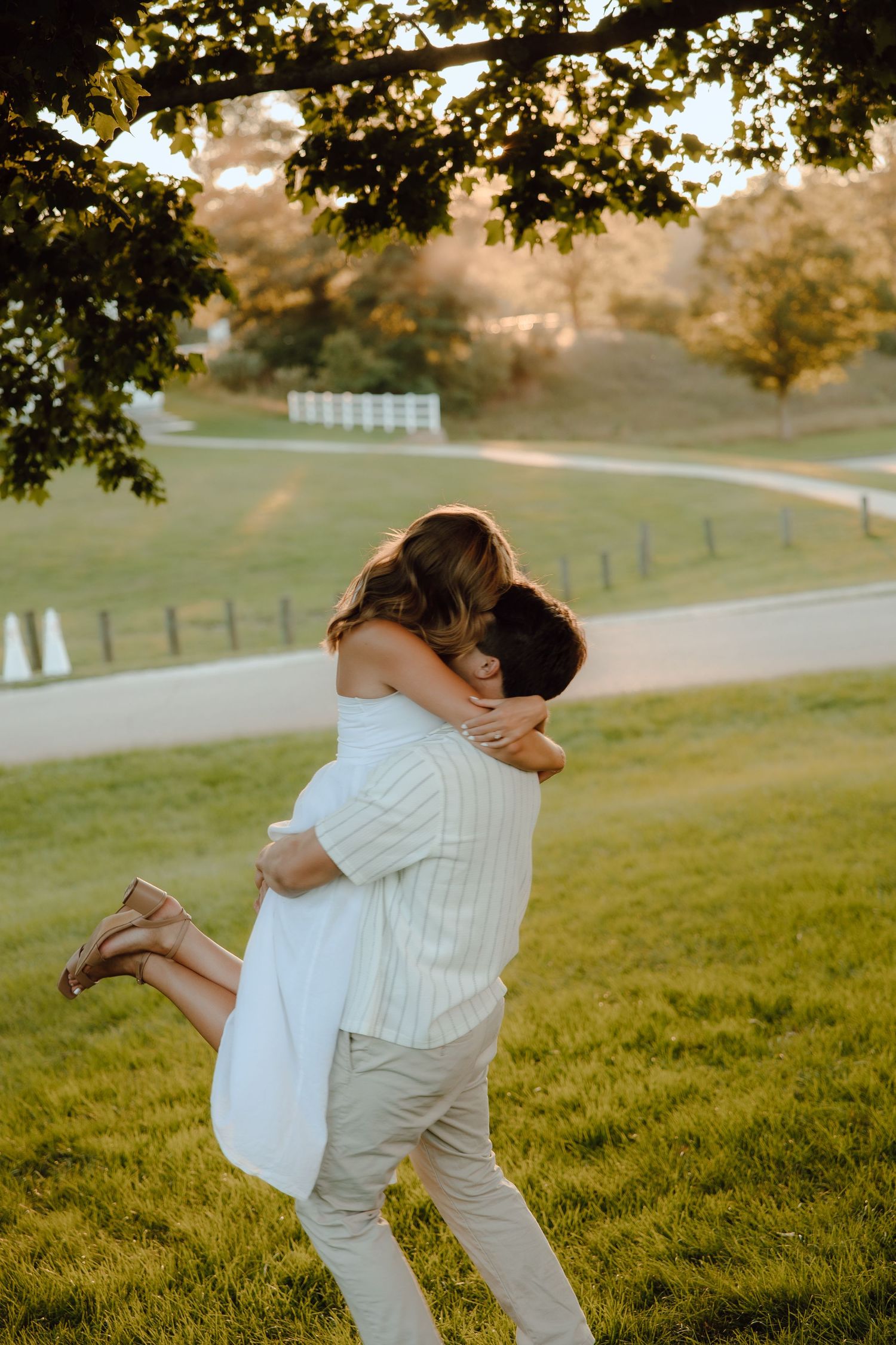 Couple sharing a romantic embrace under a tree at golden hour on a lush green lawn.