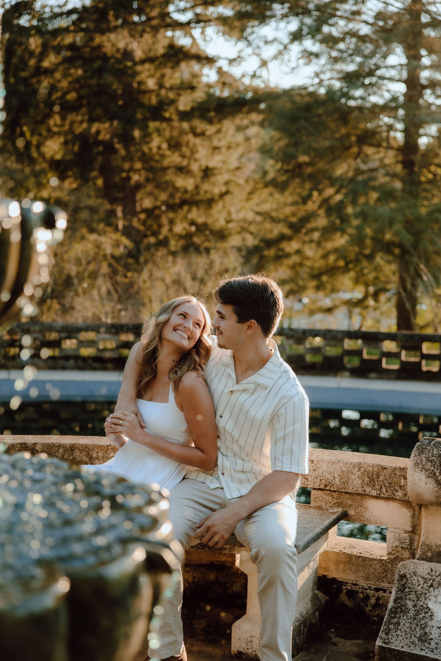 A smiling couple sits together on a stone fountain ledge surrounded by golden trees in a sunlit outdoor park.