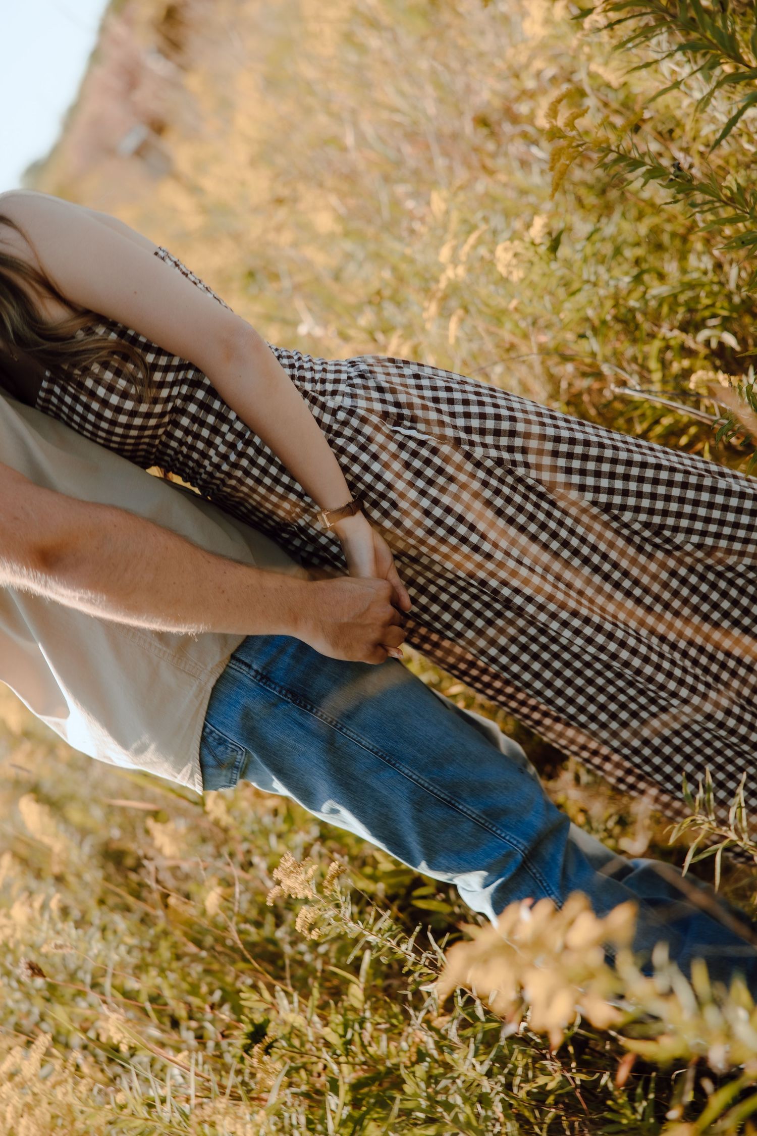 Couple embracing outdoors in golden field, woman wearing checkered midi skirt and man in blue jeans.