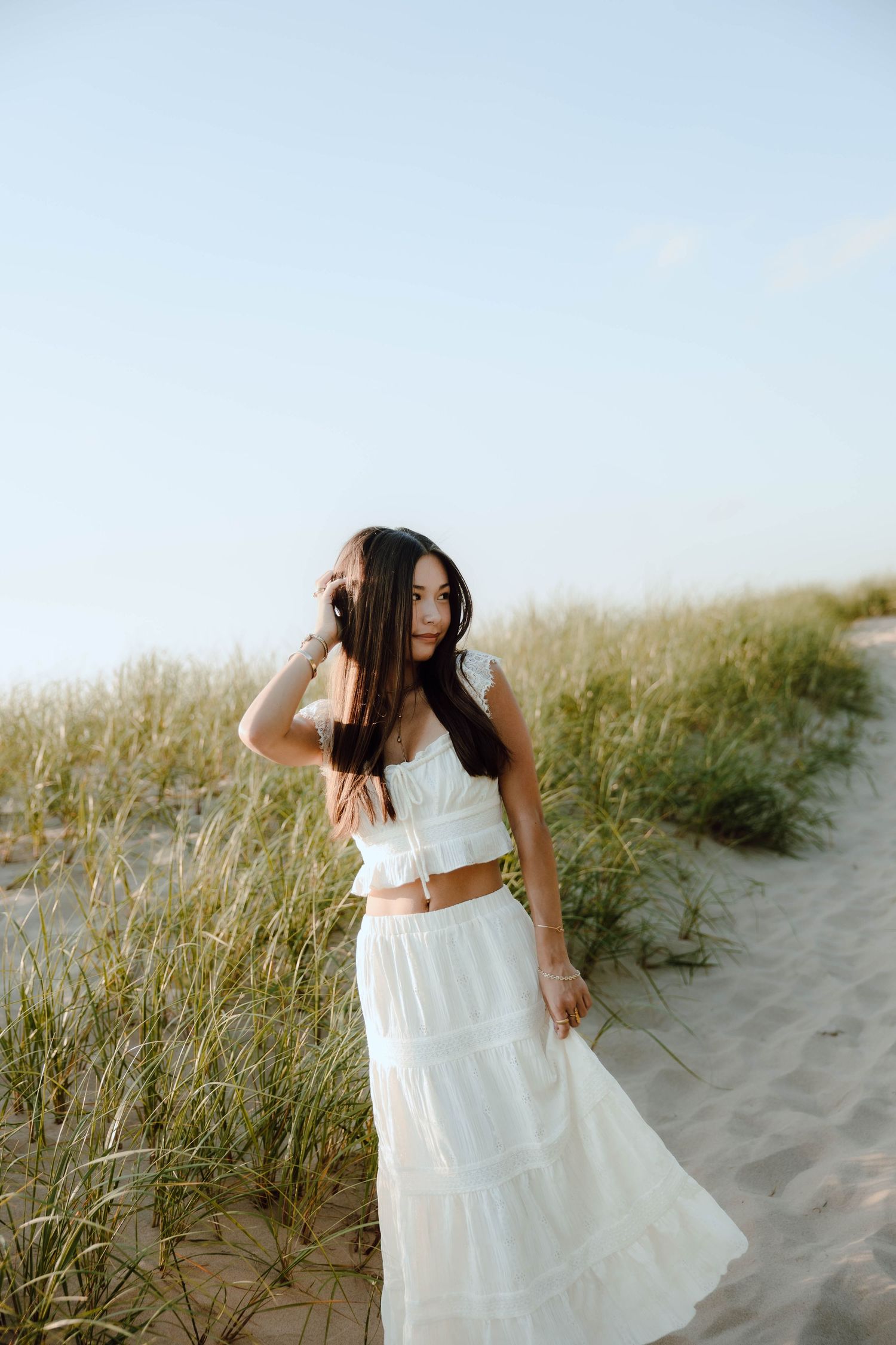 Young woman in white crop top and maxi skirt poses gracefully on sandy beach dunes with tall grass.
