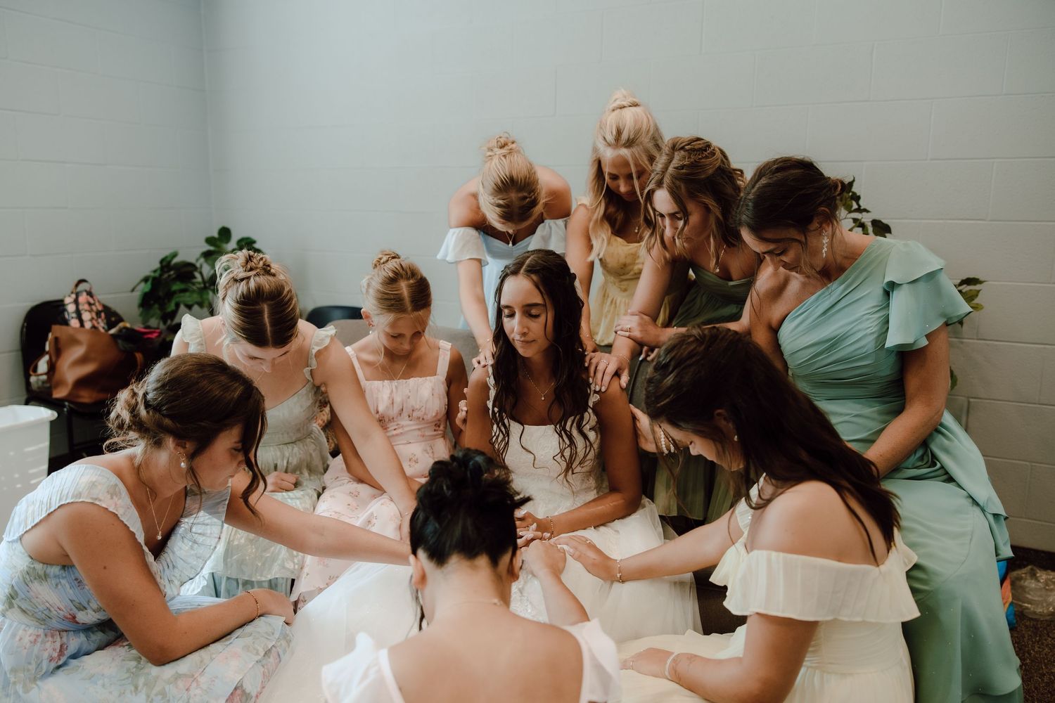 Bride surrounded by bridesmaids in colorful dresses during an intimate prayer circle indoors.