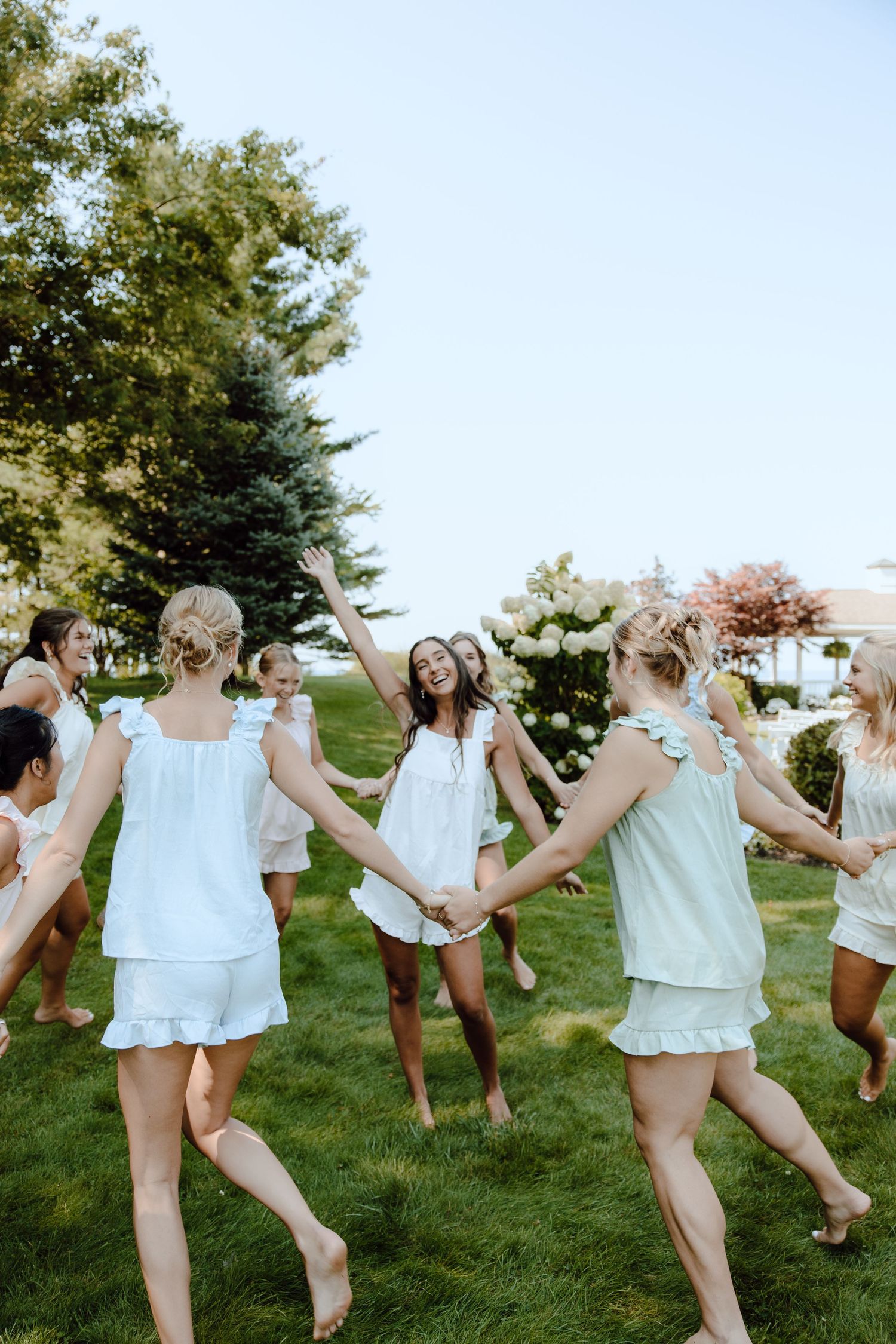 Bridesmaids in white pajamas hold hands dancing in a circle on green grass outdoors.