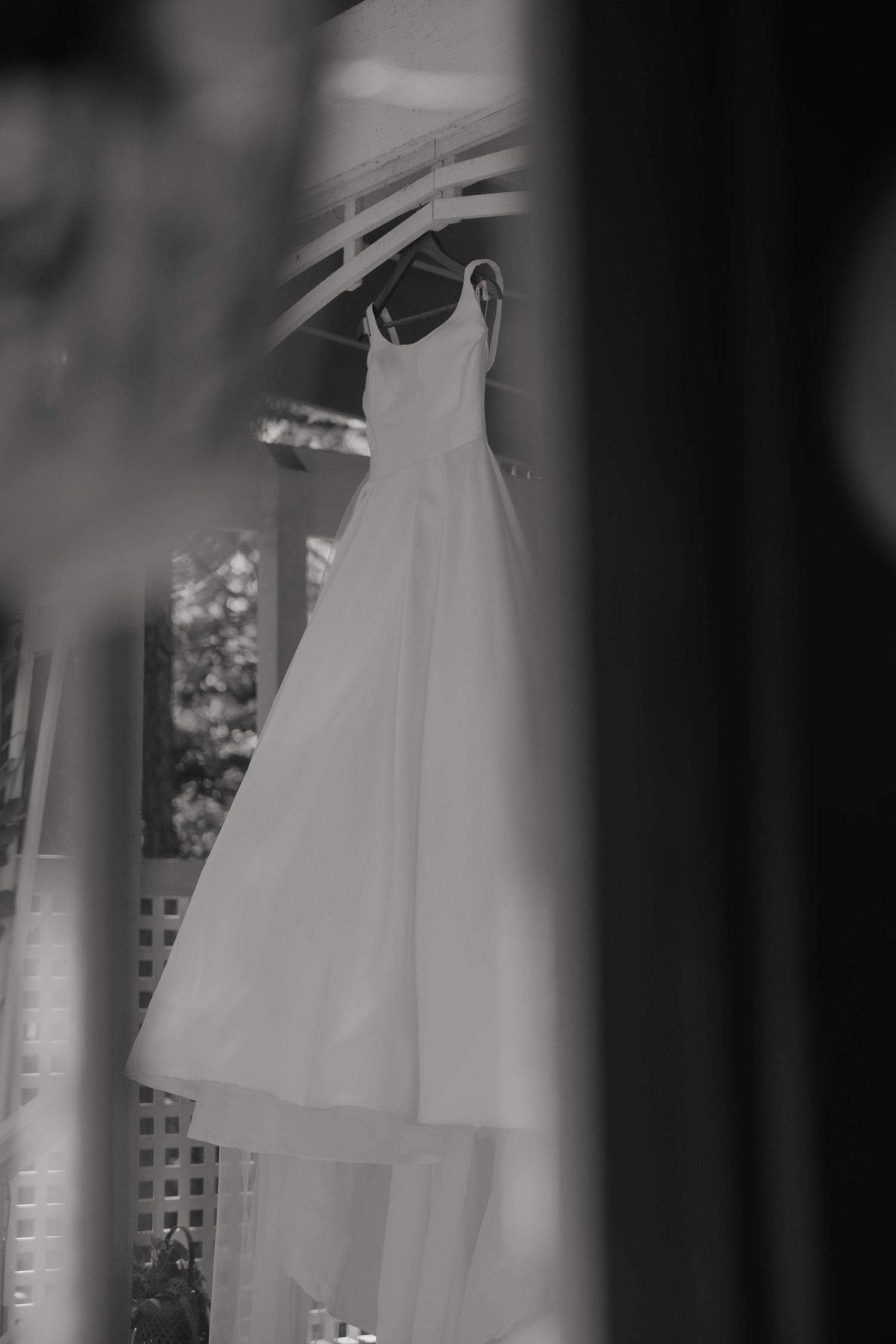 Black and white photo of an elegant wedding dress hanging on a white pergola outdoors.