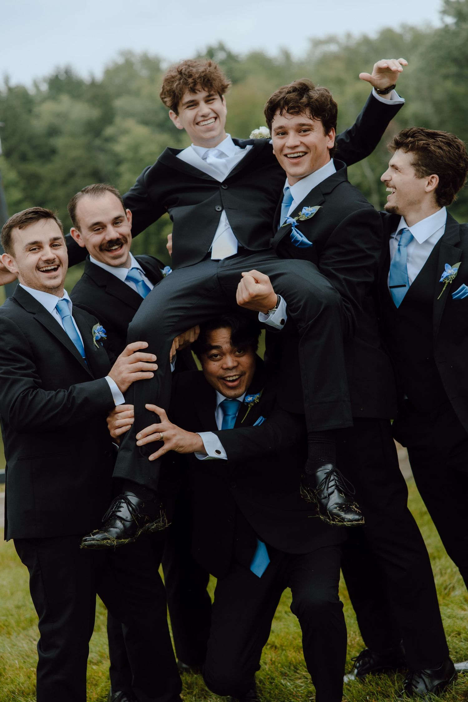 Groomsmen in black suits with blue ties lifting groom outdoors at wedding.