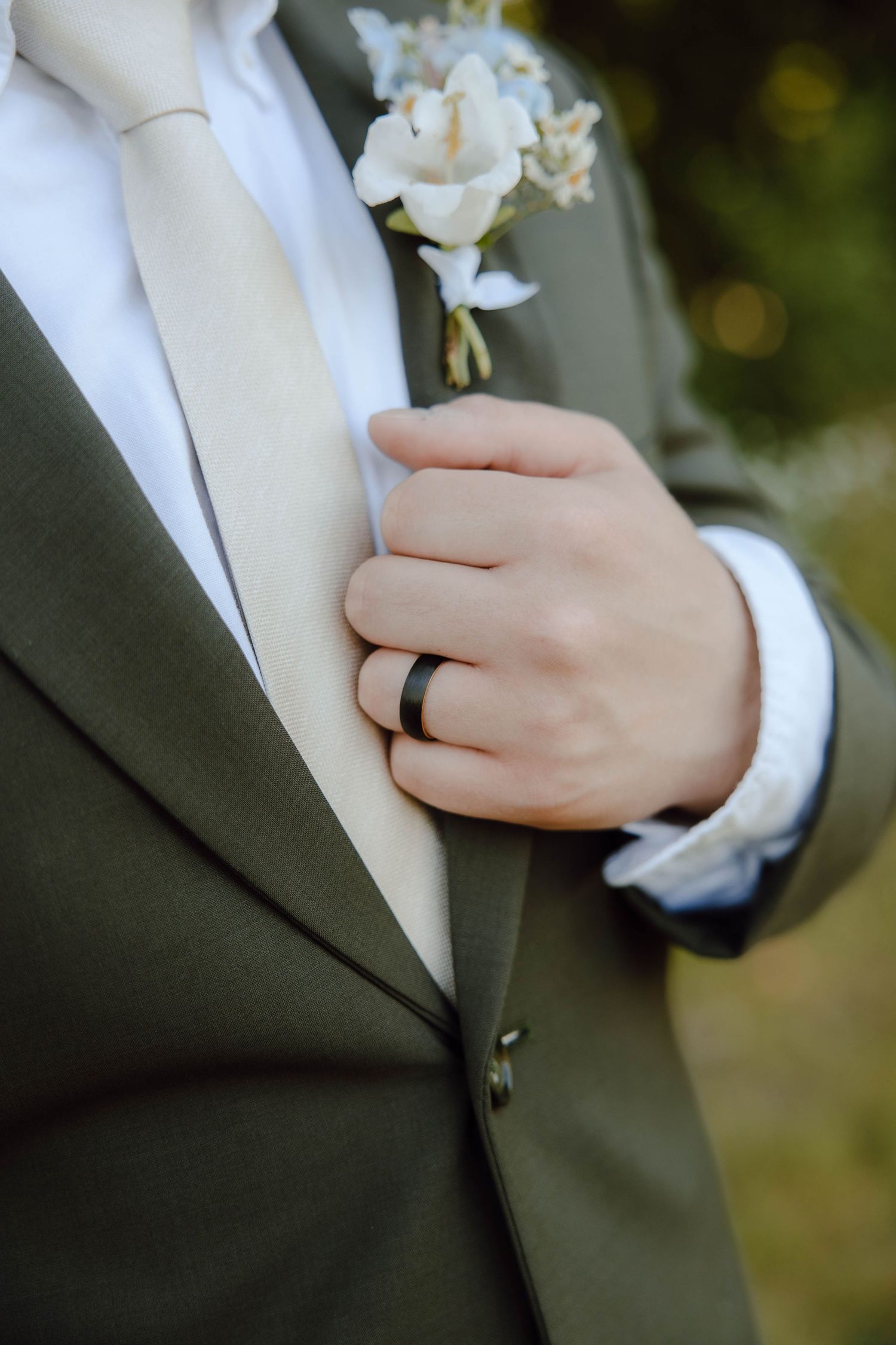 Groom in olive suit adjusting boutonniere with black wedding band visible on ring finger.