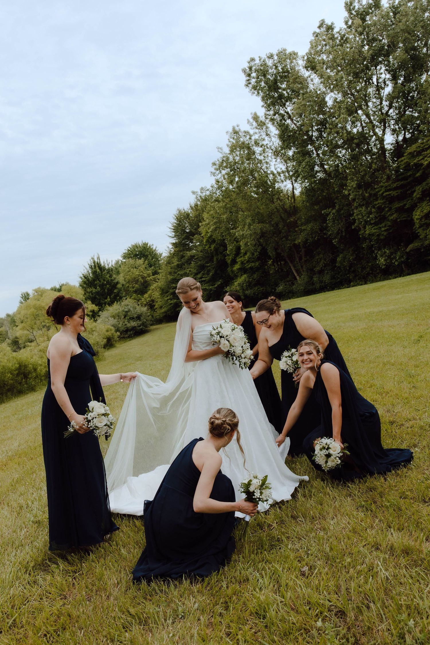 A group in navy blue dresses gathers on a grassy hillside with a white dress during a wedding photo session.