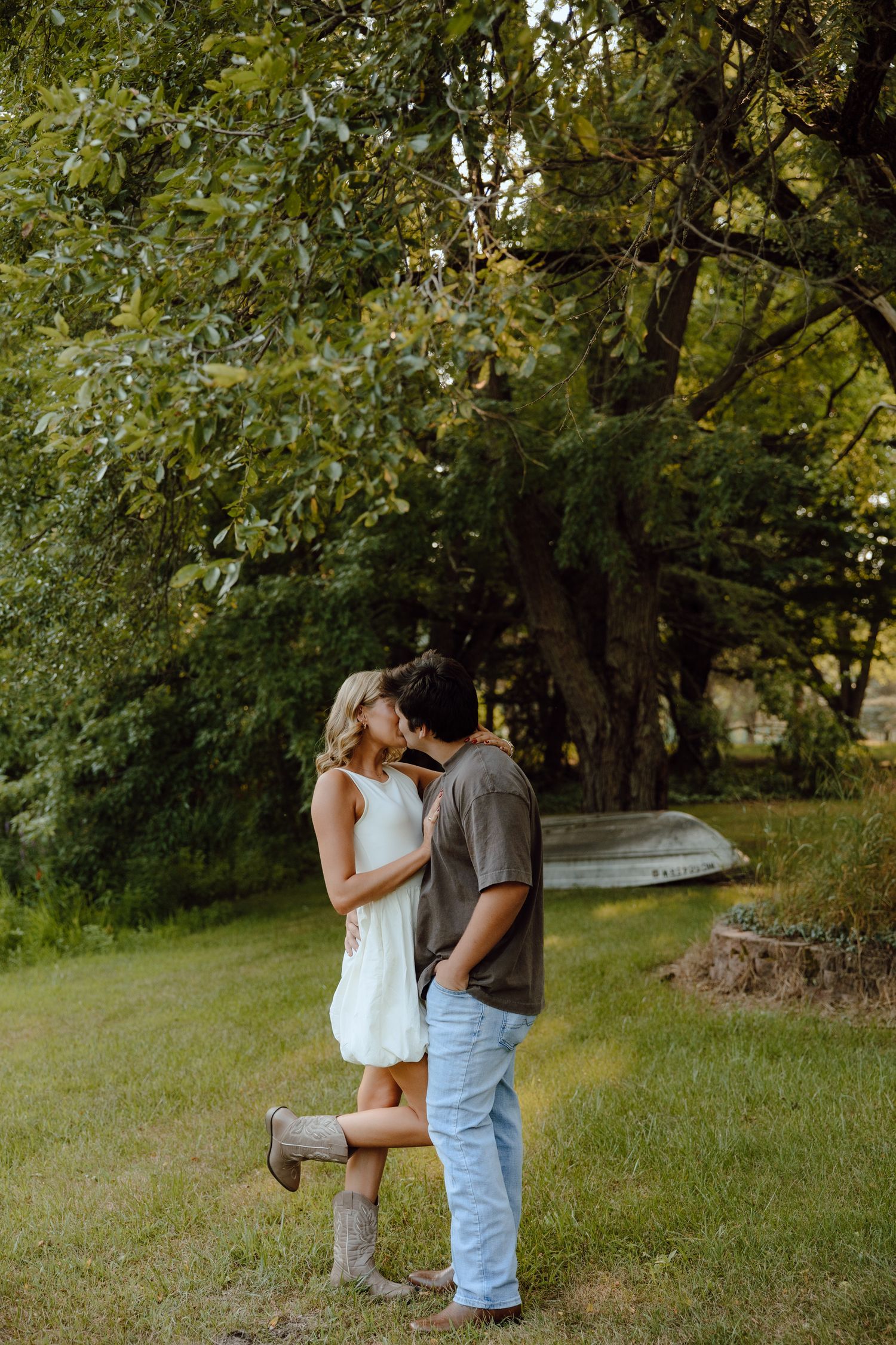 A couple embraces in a romantic pose among trees with a boat visible in the background.