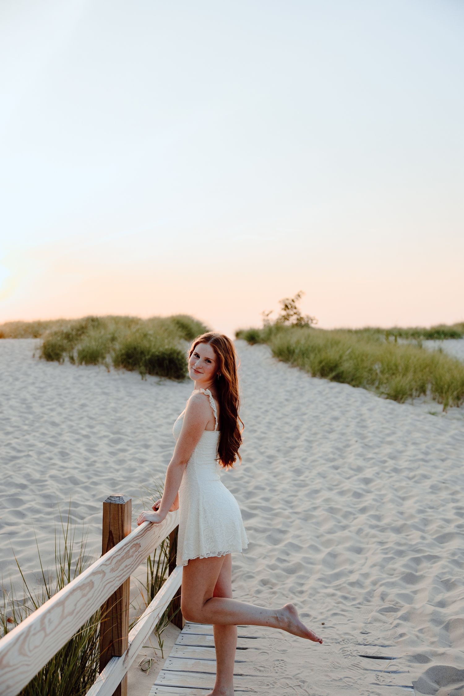 Person in white dress poses on wooden boardwalk at beach during golden hour.