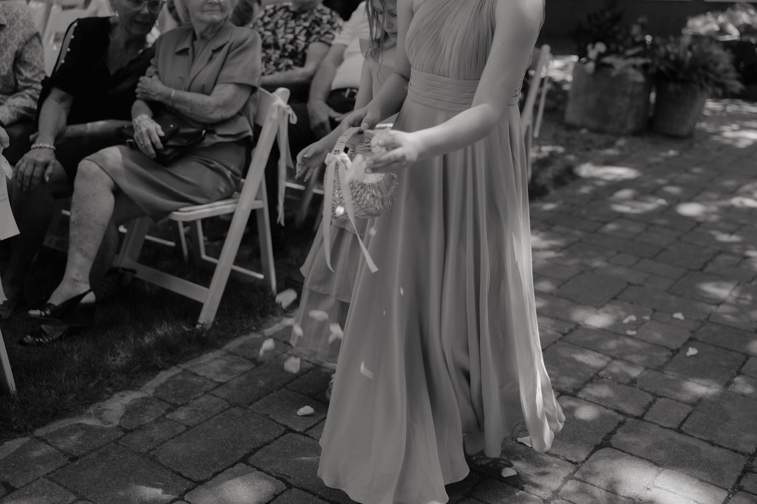 Black and white photo of a flower girl in a long dress scattering petals at an outdoor wedding ceremony.
