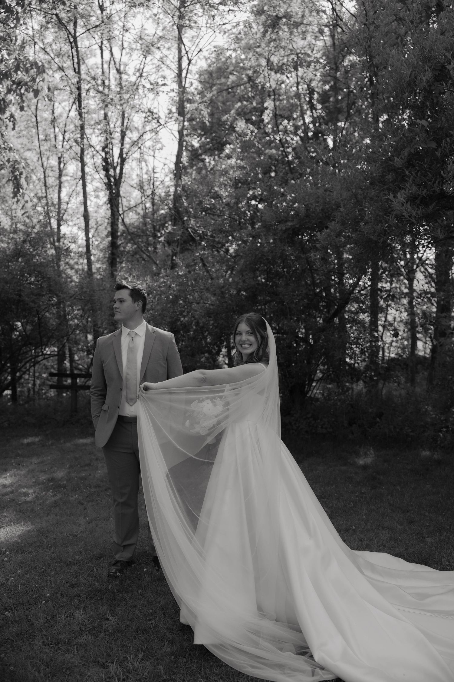 Black and white photo of a bride and groom walking outdoors, her long veil trailing behind them among trees.