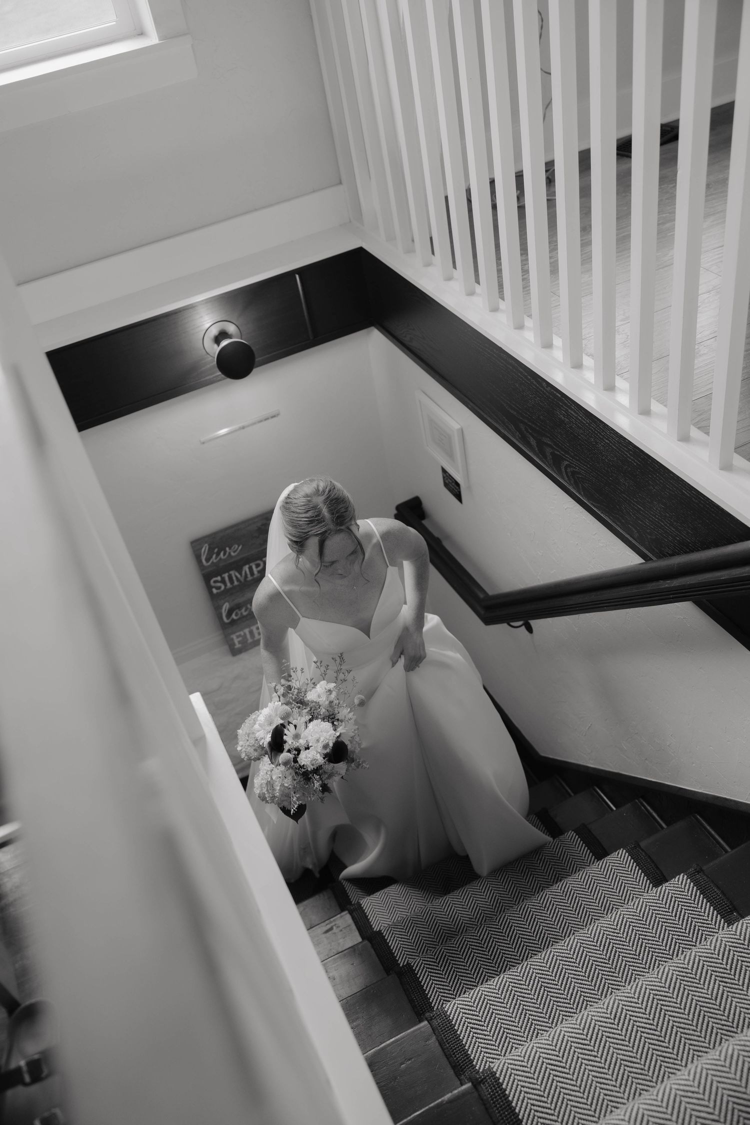 Black and white aerial view of a bride in a white gown holding a bouquet while descending an elegant staircase.