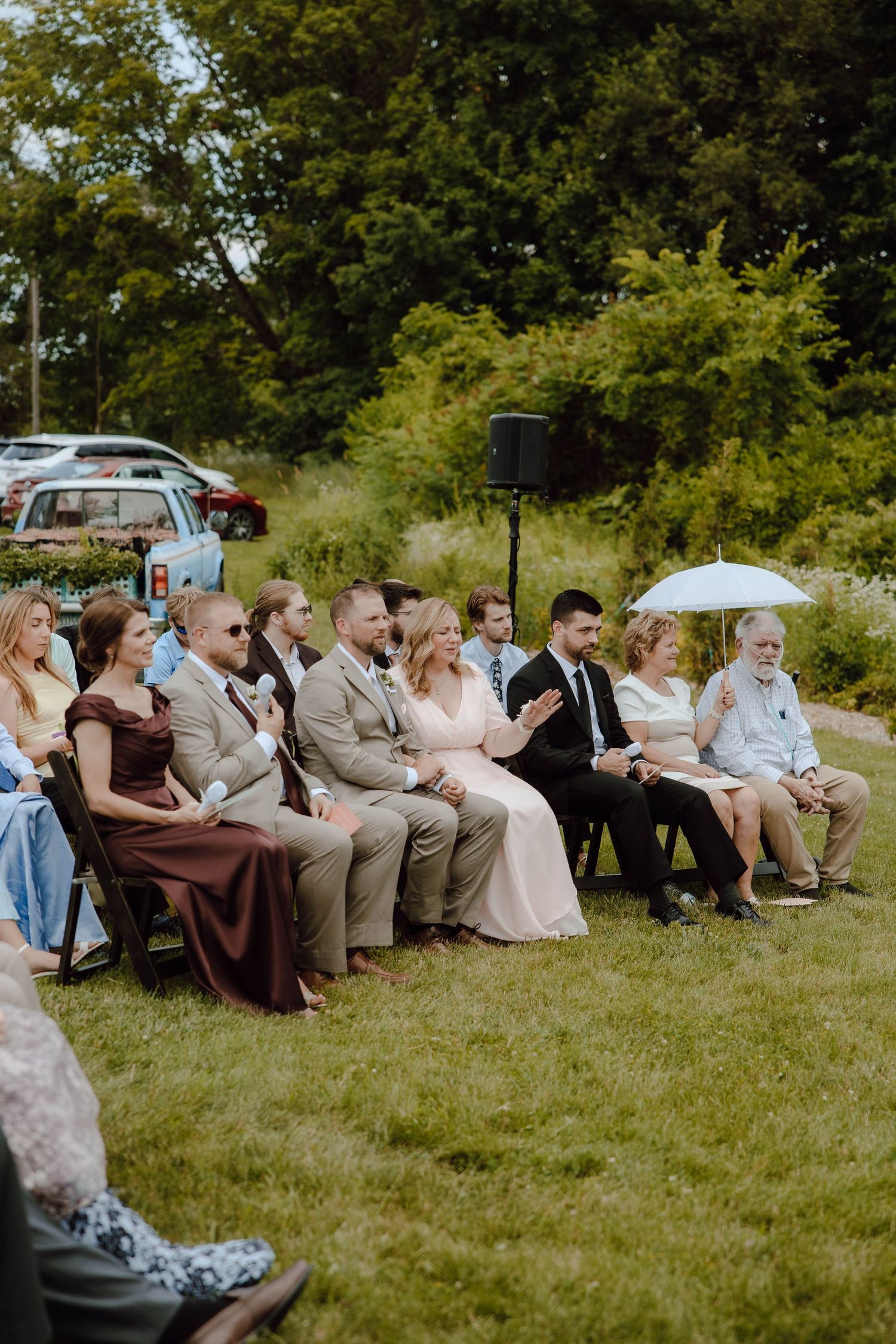 Wedding guests seated in wooden chairs on green lawn during an outdoor ceremony on a sunny day.