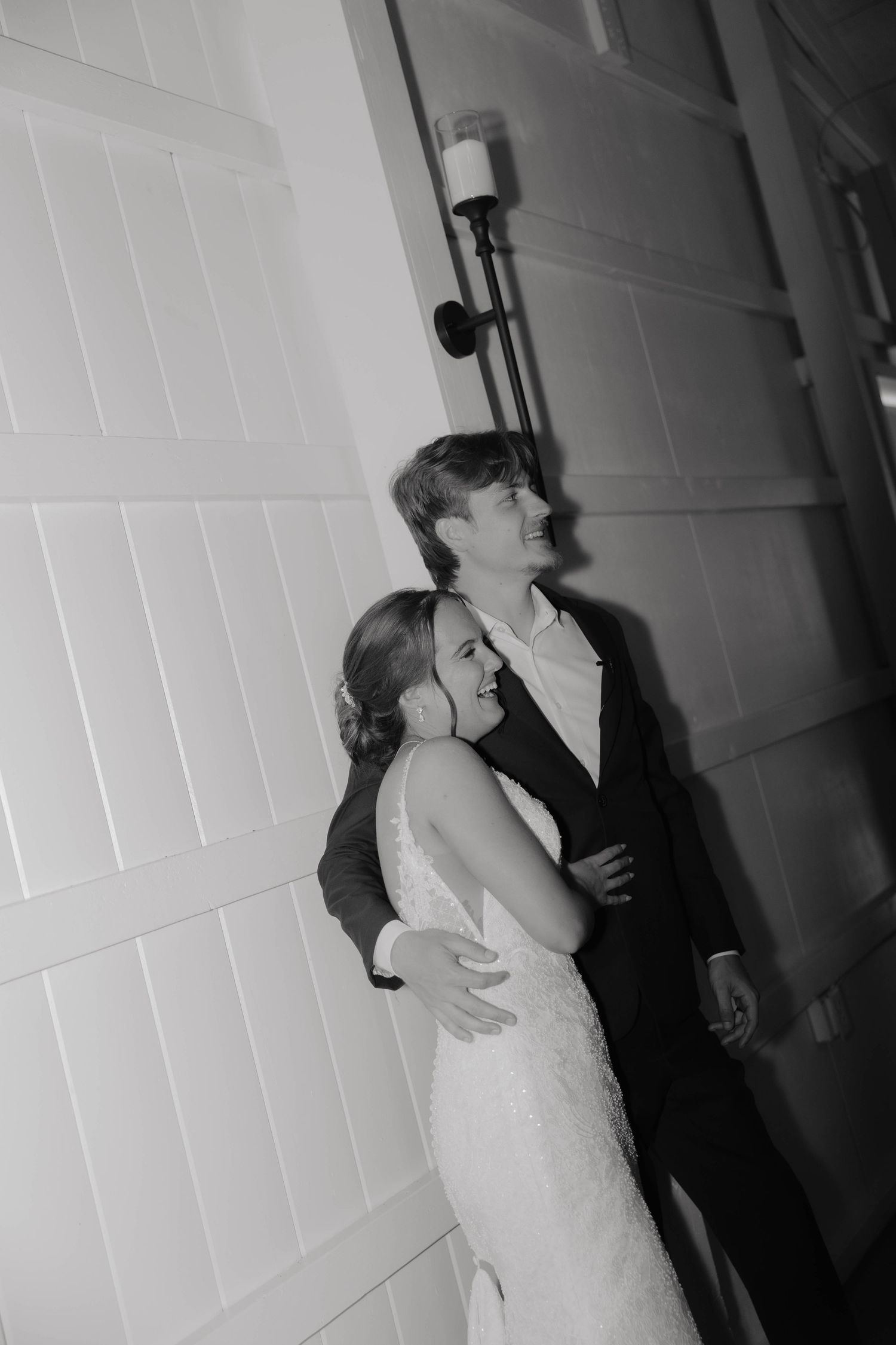 Black and white photo of a smiling bride and groom embracing against a white wall with a candle sconce.