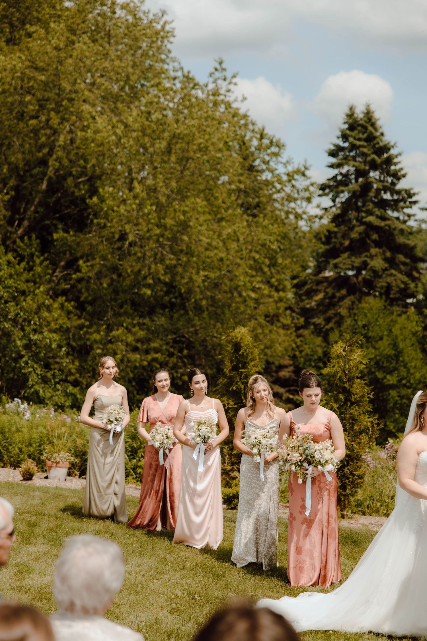 Five bridesmaids in mismatched blush and gold gowns holding white bouquets stand outdoors during a garden wedding ceremony.