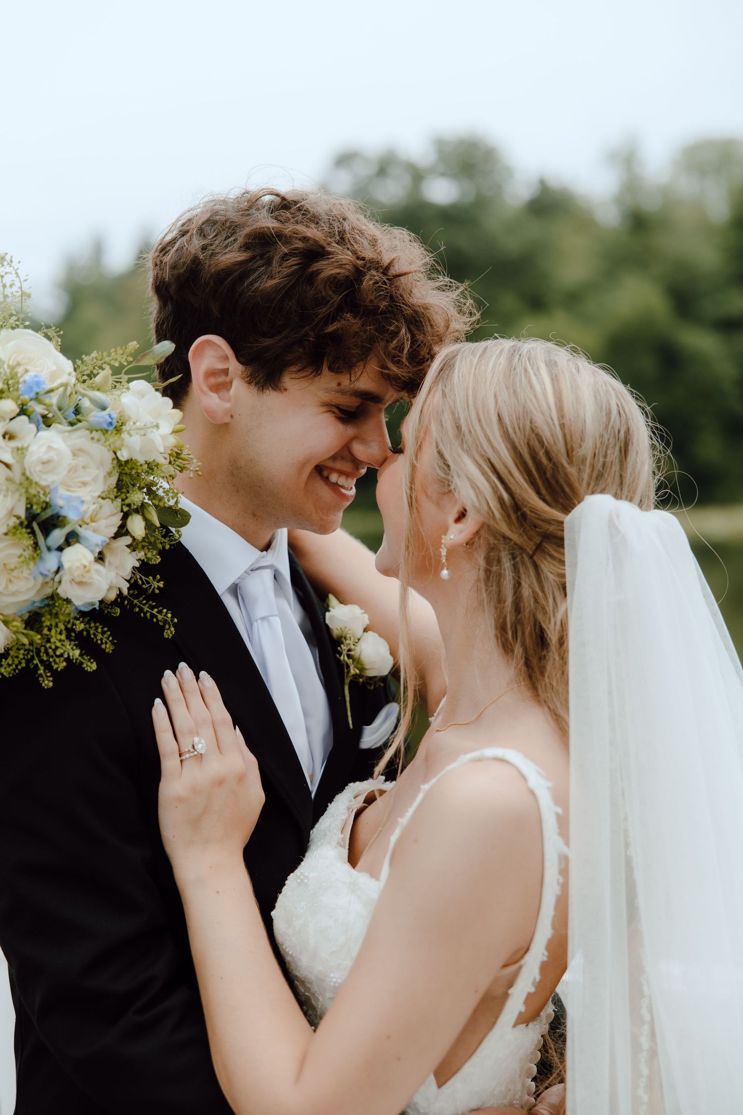 Smiling bride and groom touching foreheads holding white floral bouquet outdoors.