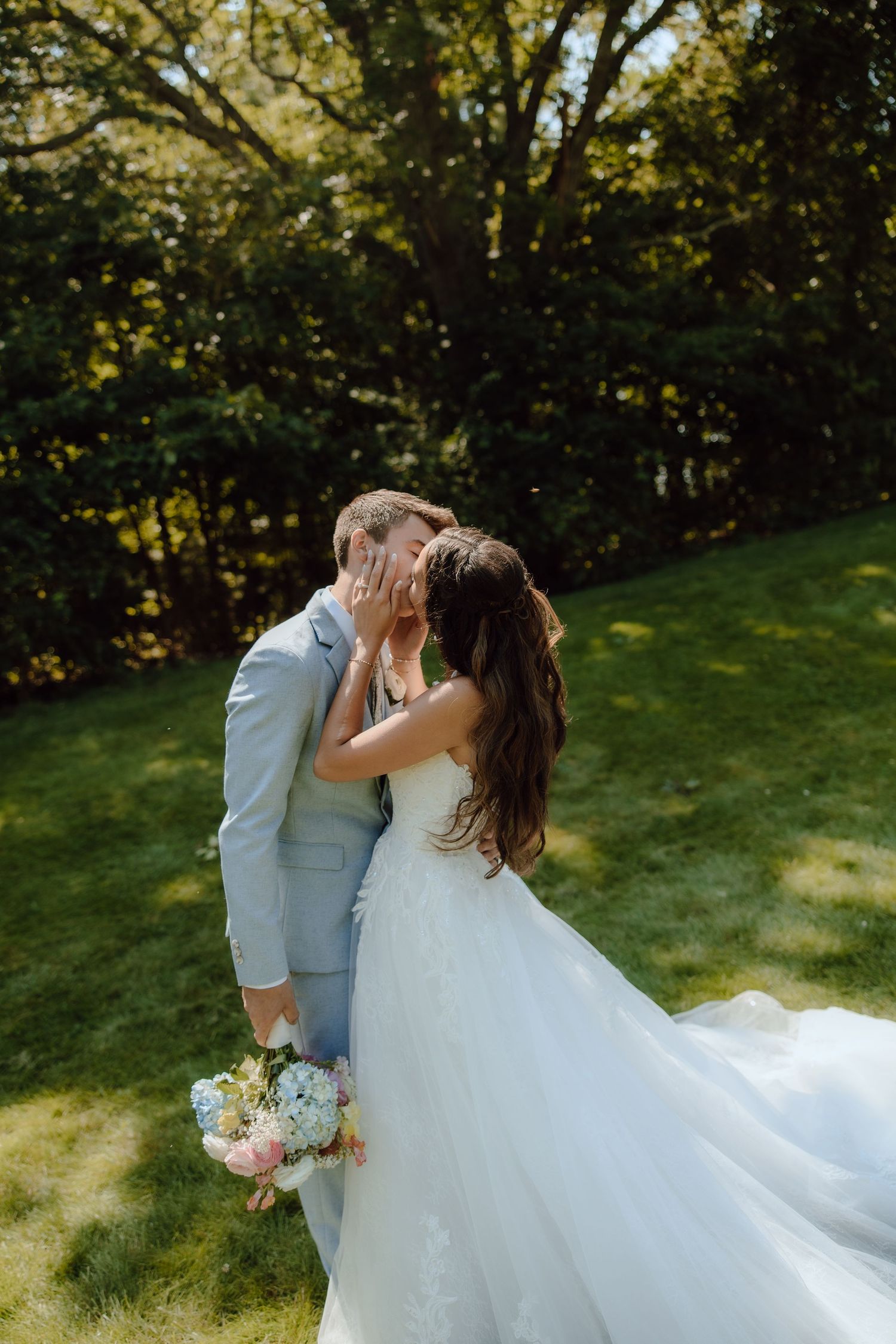A bride and groom share a romantic kiss outdoors on a sunny day, with the bride holding a pastel bouquet in a flowing white gown.