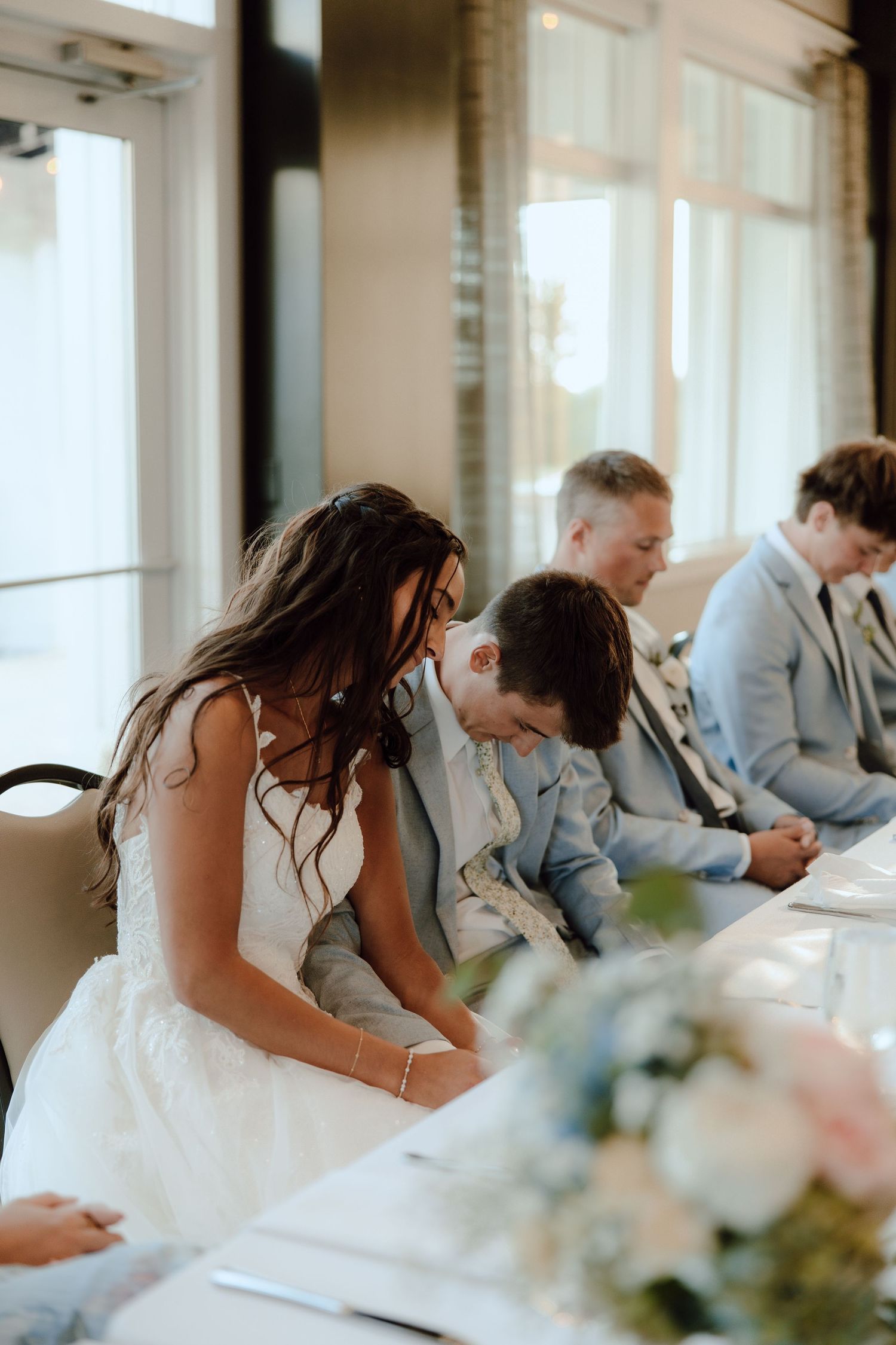 Bride leaning toward young guest at wedding reception table adorned with floral centerpiece.