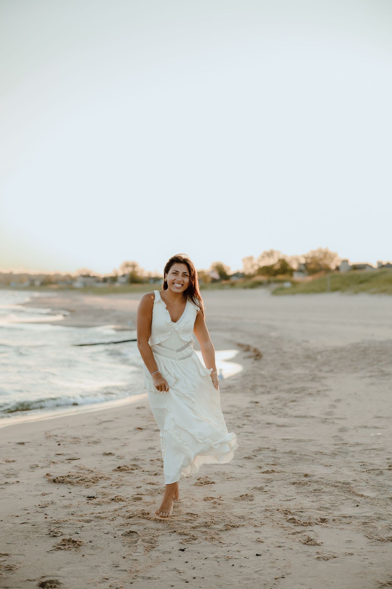 Woman in white dress walking barefoot on sandy beach at sunset, smiling joyfully with ocean waves in background.