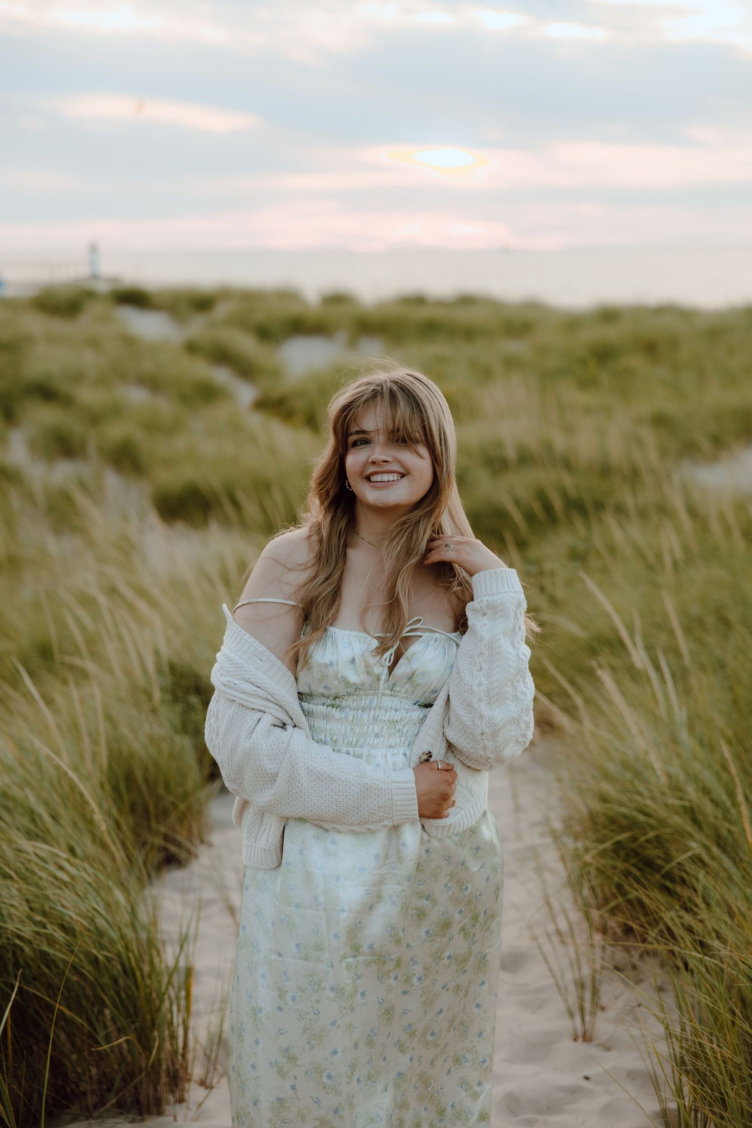 Young woman in floral dress and white jacket smiling on sandy beach path surrounded by tall grass at sunset.