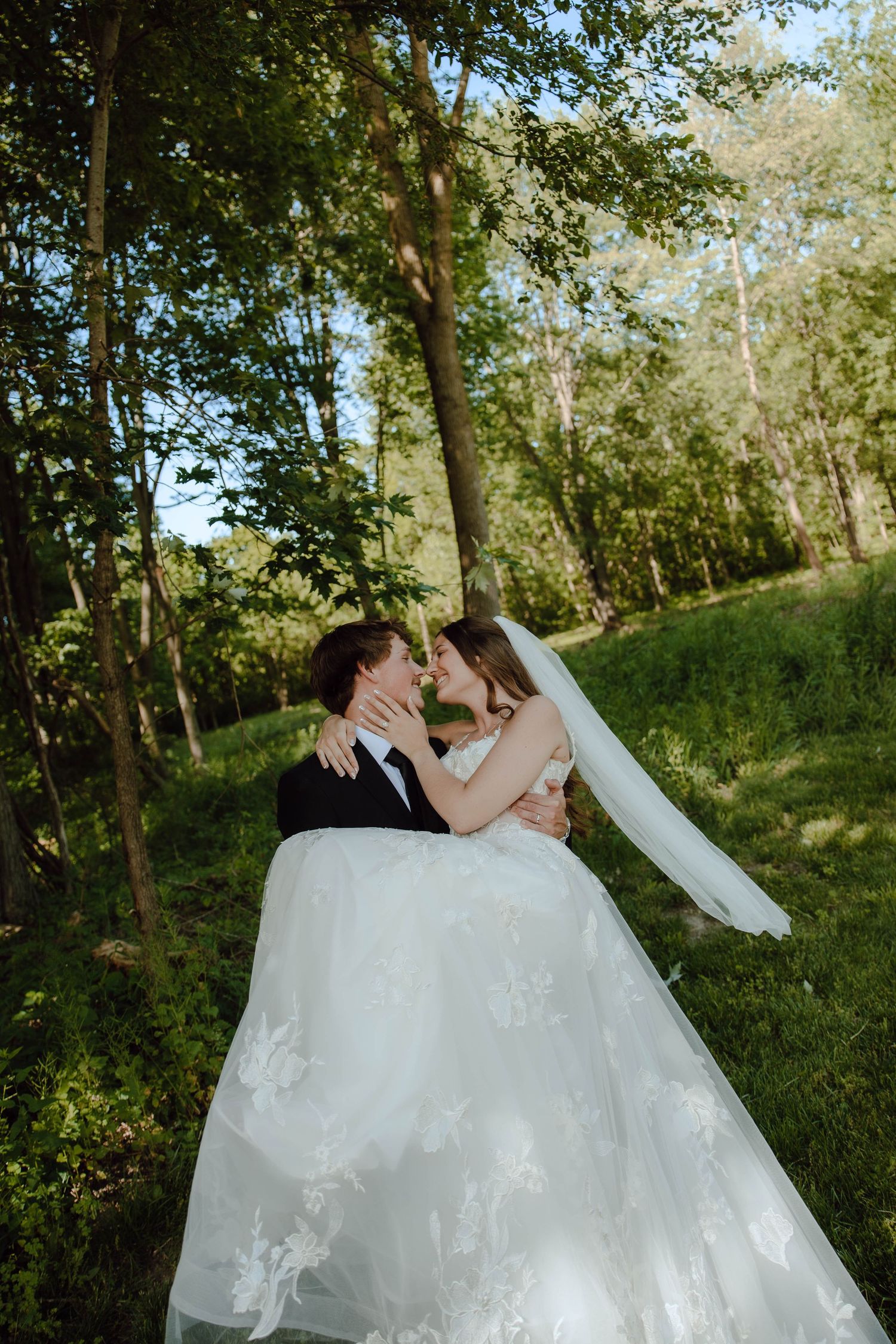 Groom carrying bride in white lace gown and veil in a lush green forest during a romantic outdoor wedding.