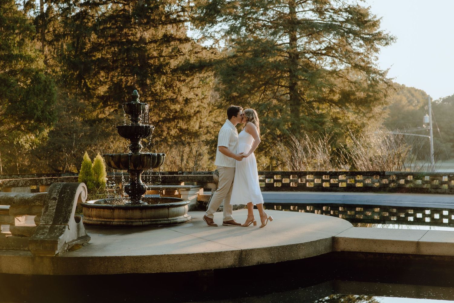 Couple embracing by a stone fountain at golden hour with lush trees and a serene pond in the background.