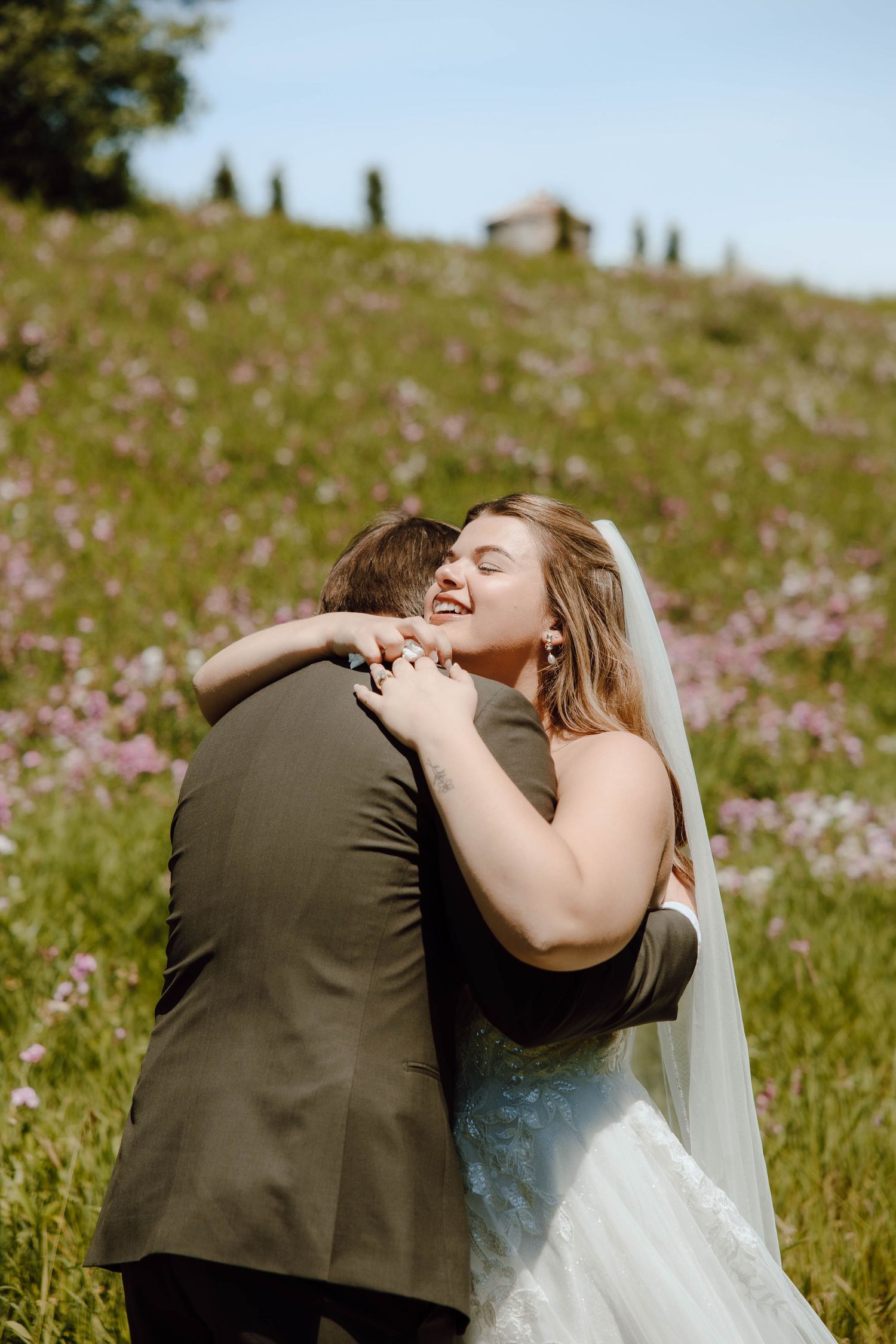 Bride and groom embracing in a wildflower meadow, bride smiling joyfully in white gown and veil on sunny wedding day.