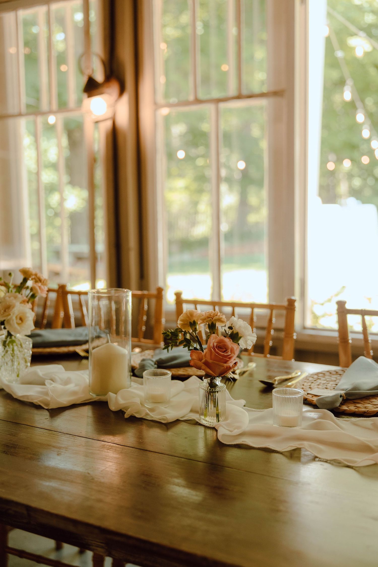 Elegant rustic wedding reception table with white floral centerpieces, candles, and gauze runner in a sunlit venue.