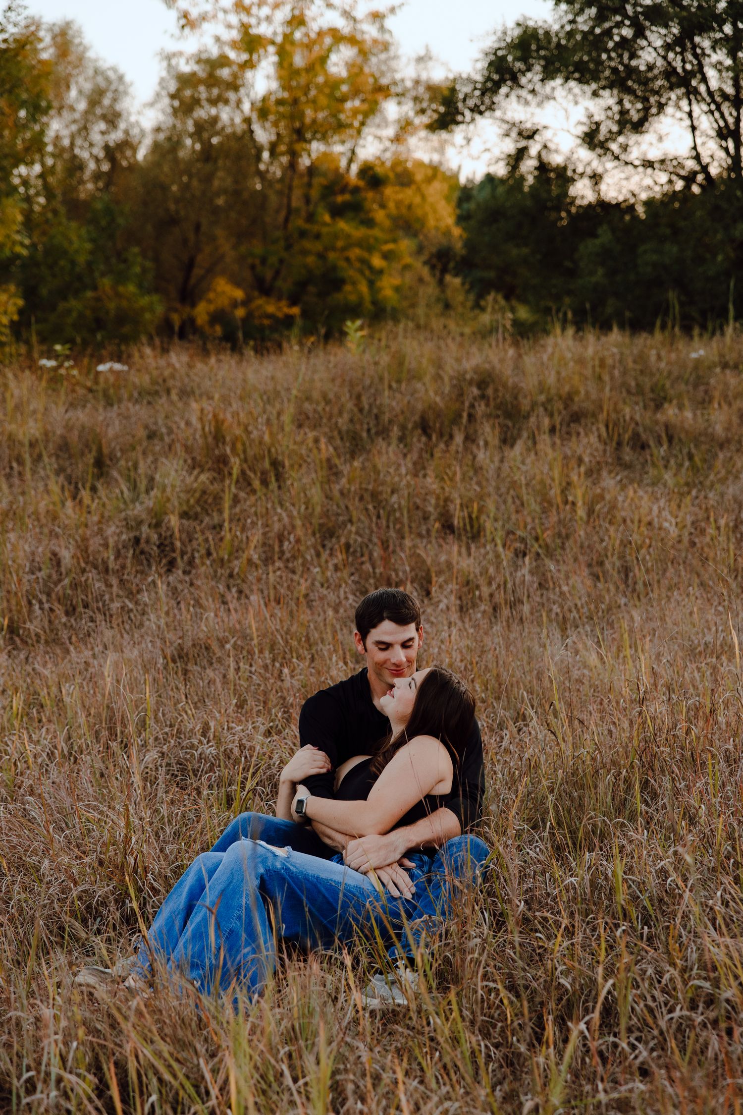 Person in black shirt and blue jeans sitting in tall autumn grass at sunset.