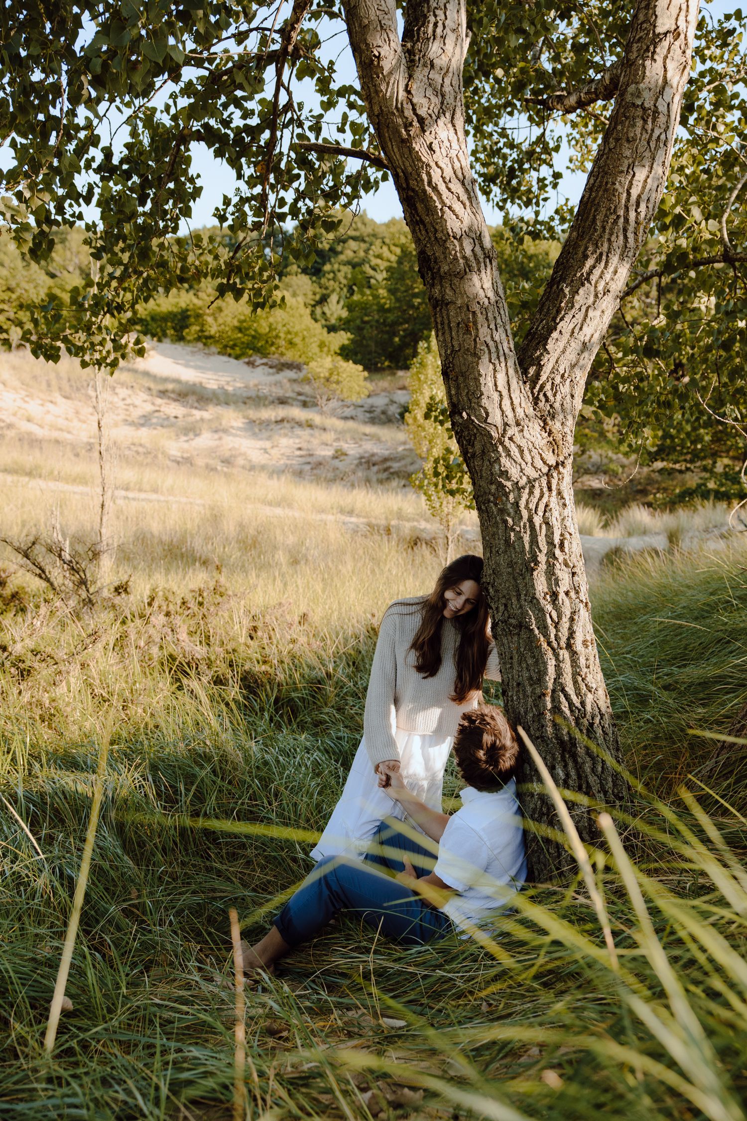 A couple sits together under a large tree in a grassy field on a sunny day.
