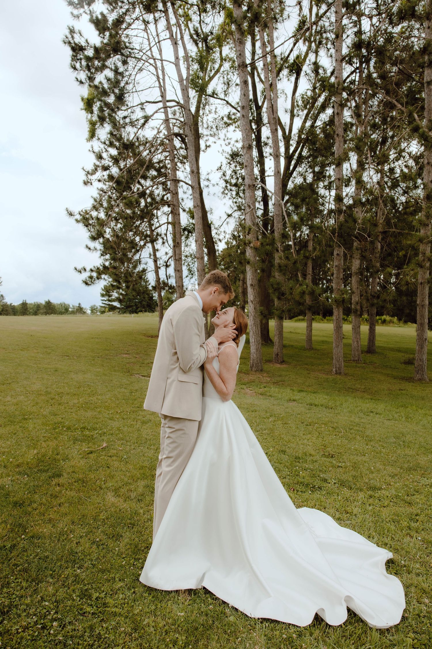 Bride in white gown and groom in beige suit share a romantic embrace on green grass surrounded by tall trees.