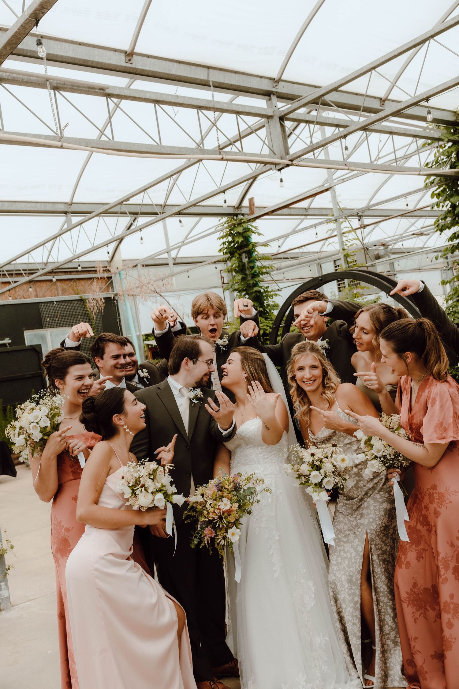 Wedding party celebrating with bride and groom in a greenhouse venue, bridesmaids in pink dresses holding bouquets.