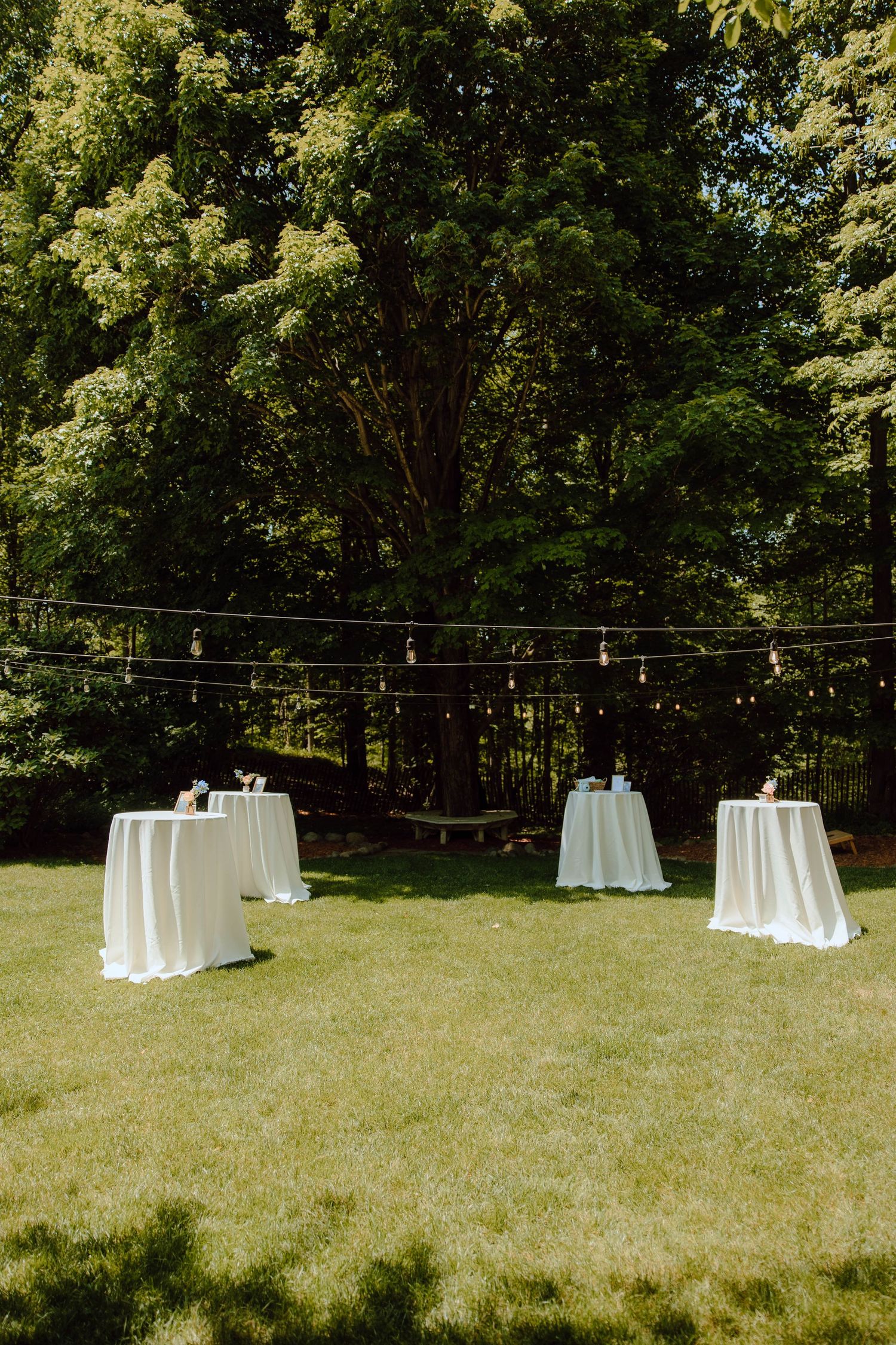Outdoor wedding cocktail hour with white draped high-top tables on green lawn surrounded by lush trees and string lights.