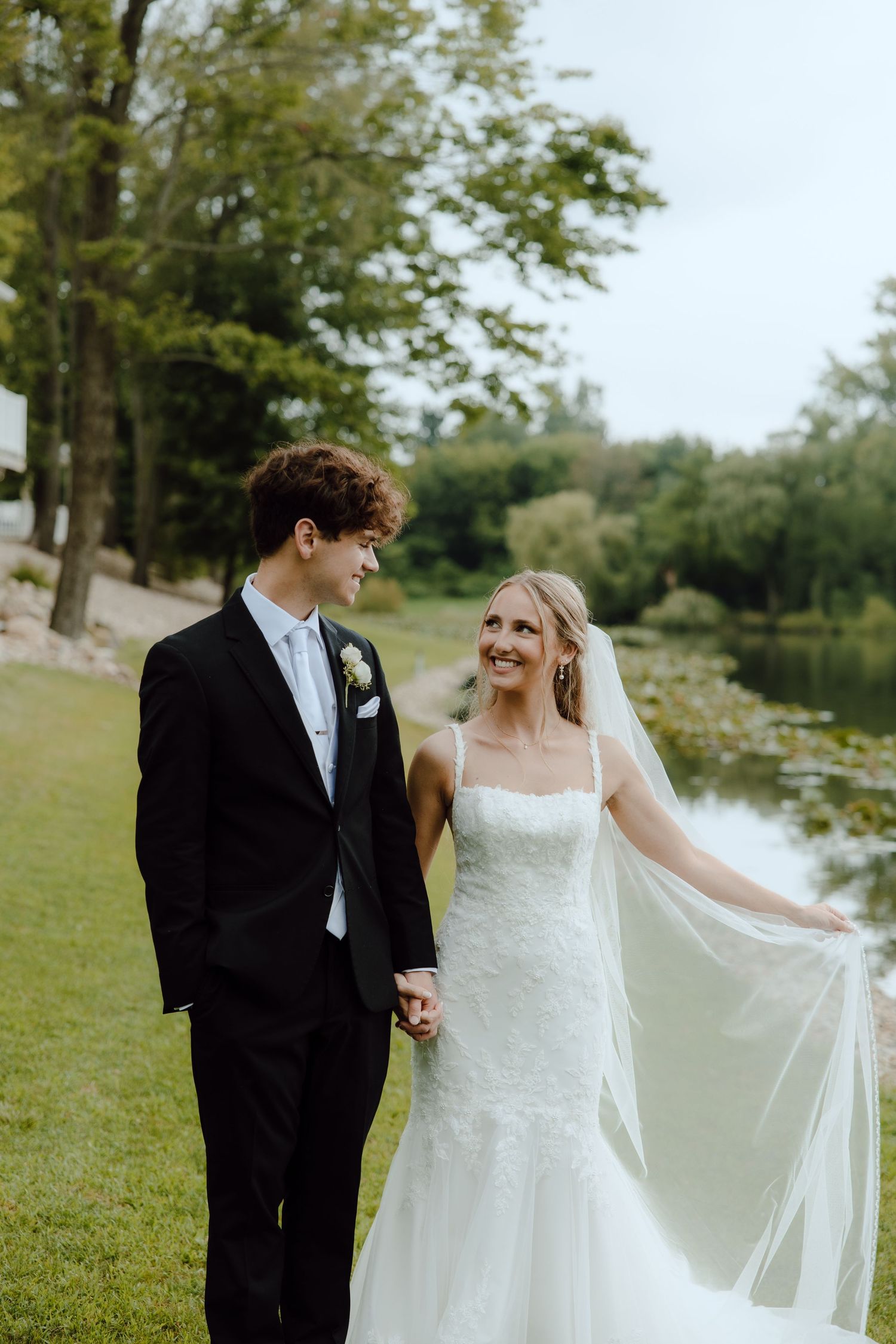 Bride and groom hold hands walking by a pond, bride in lace gown with veil, groom in black tuxedo.