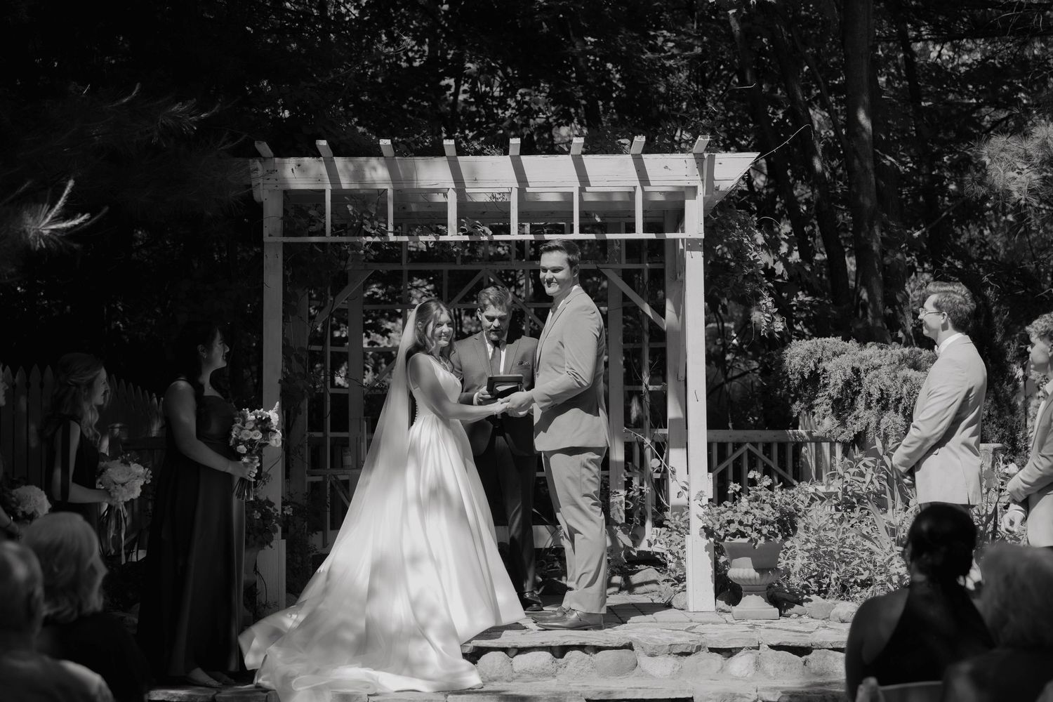 Bride and groom exchange vows at an outdoor wedding ceremony beneath a white pergola with guests seated around them.
