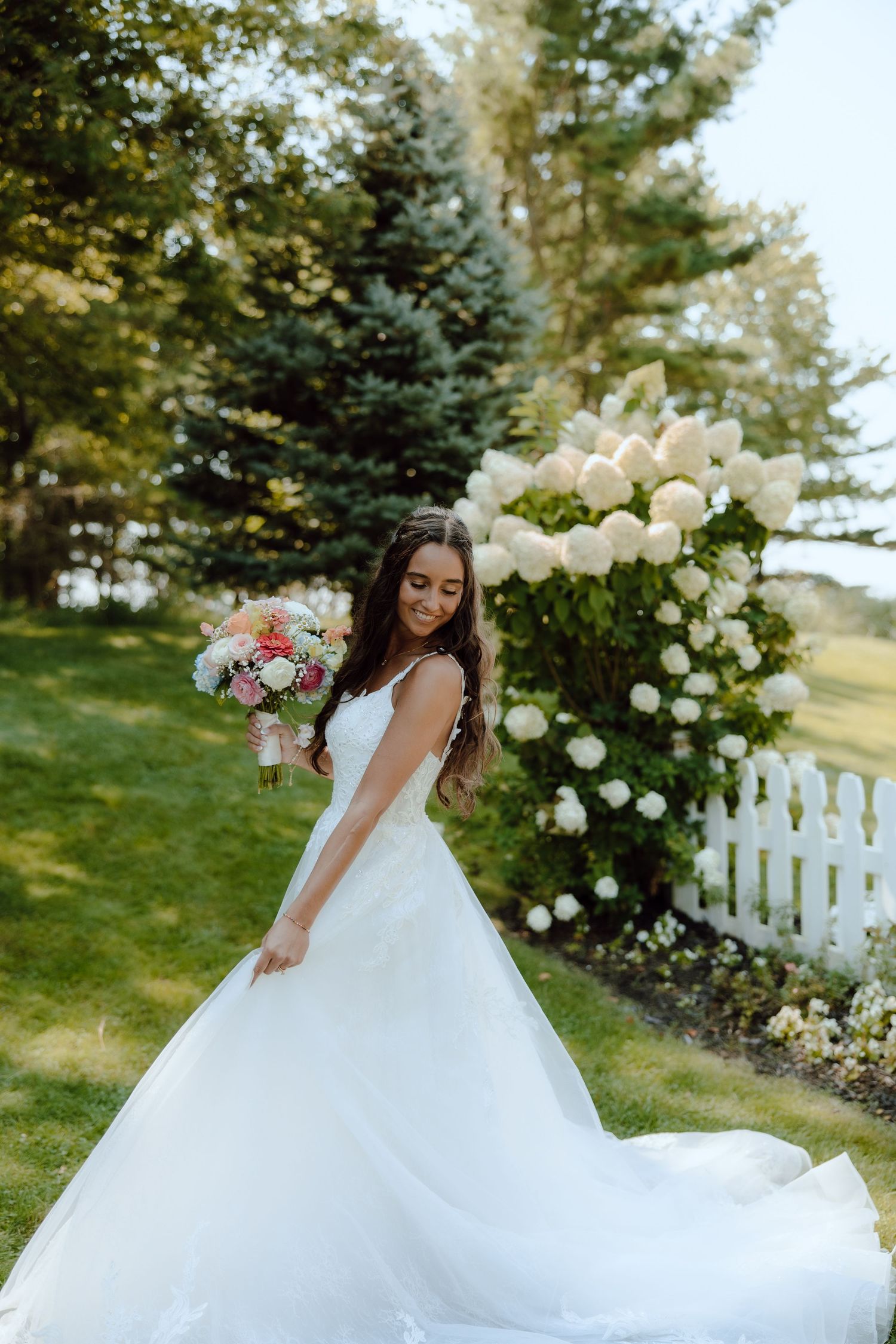 Bride in white ball gown holding bouquet beside blooming white hydrangeas in lush garden setting.