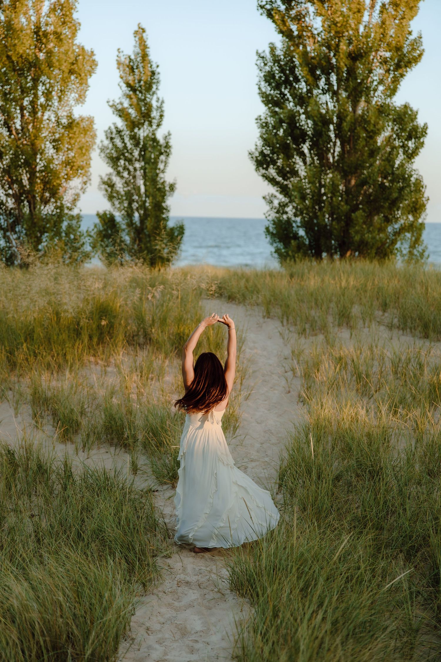 Woman in white dress dancing on sandy path between green dunes with trees and ocean in background at sunset.