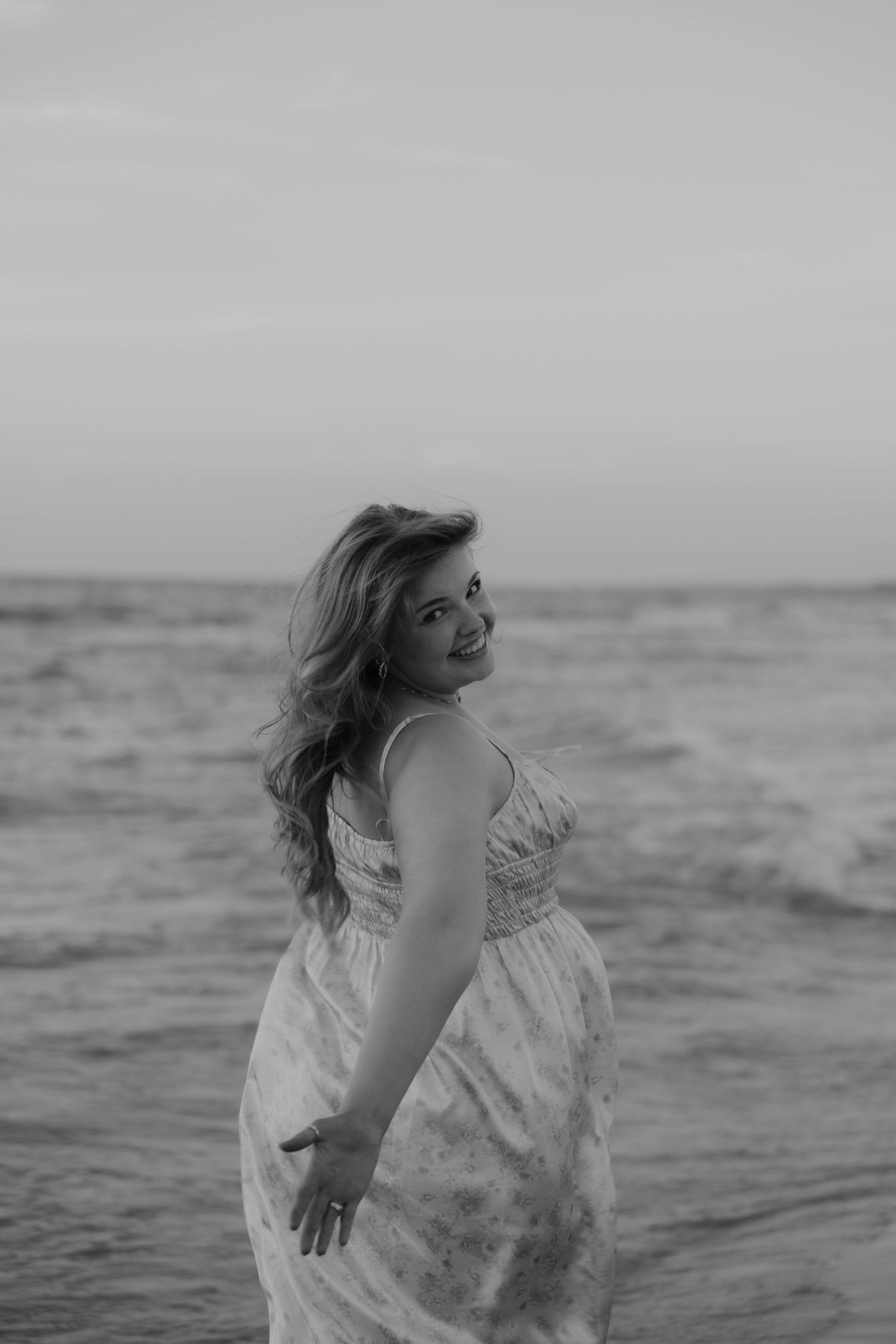 Black and white photo of a smiling woman in a flowy dress posing on a beach with ocean waves behind her.