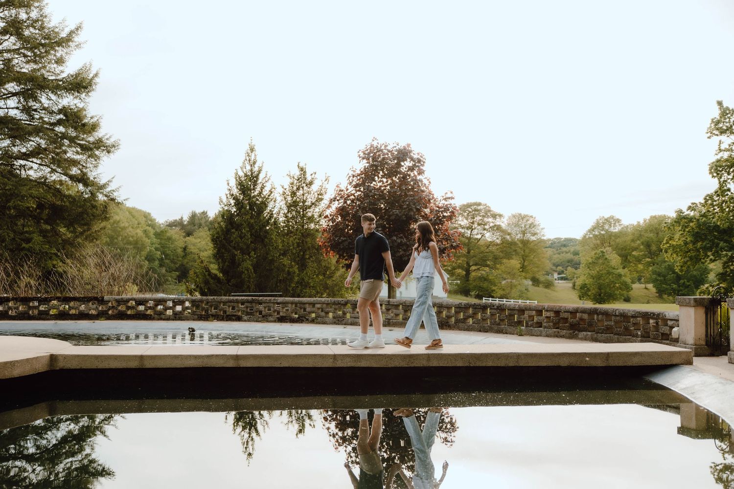 Couple holding hands by a reflective pool surrounded by lush greenery and trees in a scenic garden setting.