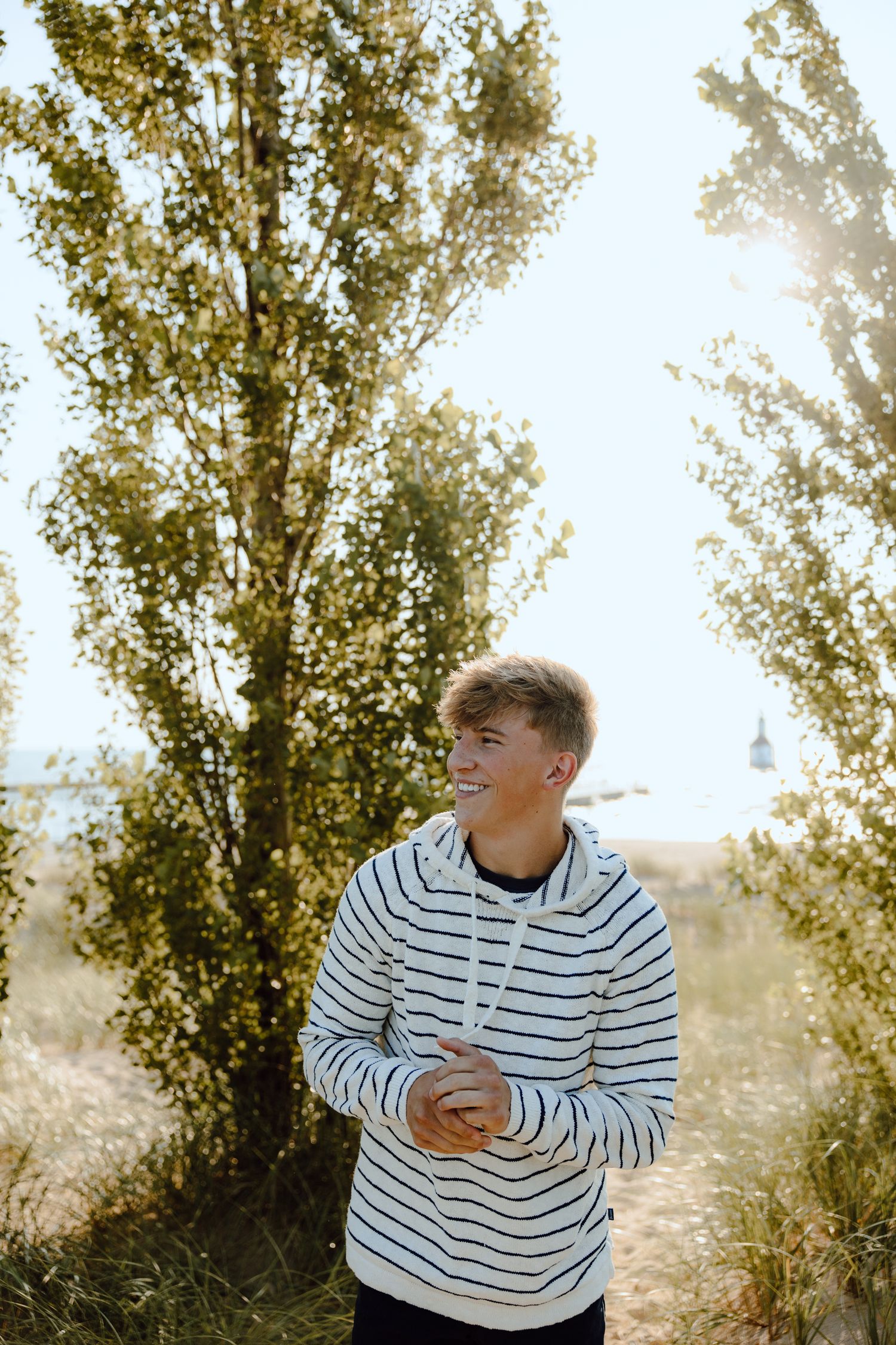 Person in striped sweater smiling outdoors with sunlit trees in background.
