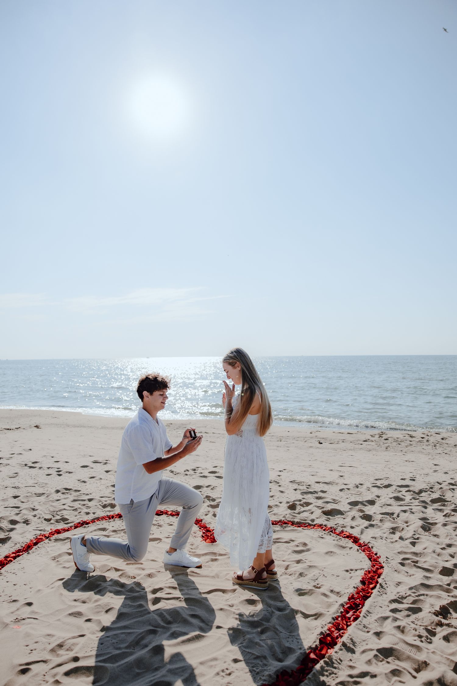 Beach proposal scene with red rose petals arranged in a heart shape on the sand.