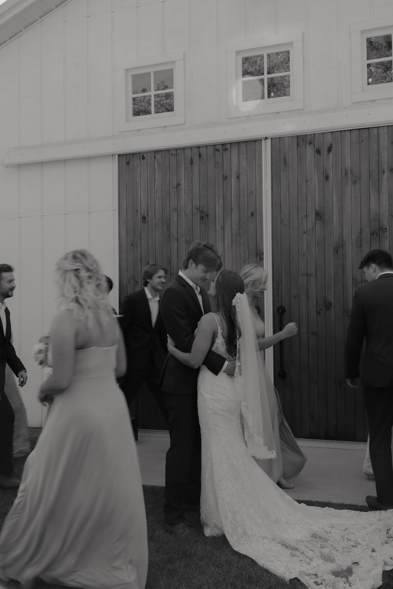 Black and white photo of bride and groom kissing outside a white barn with wedding party.