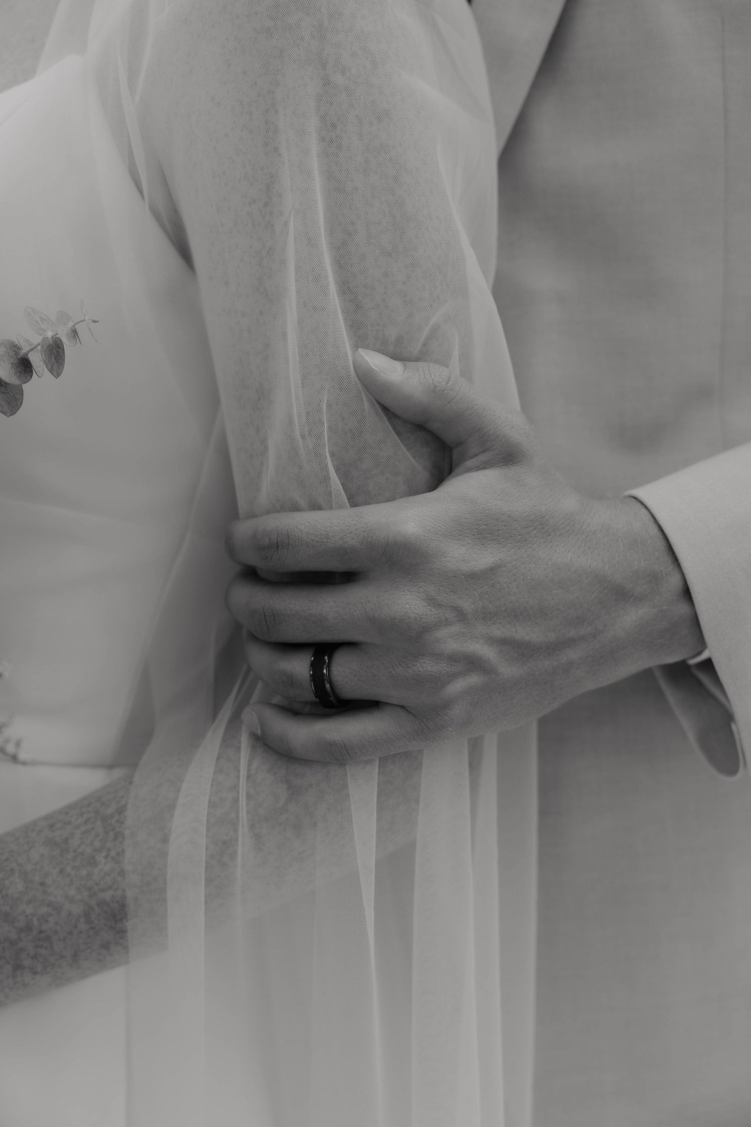 Black and white wedding photo of groom's hand with ring embracing bride in flowing white veil and gown.