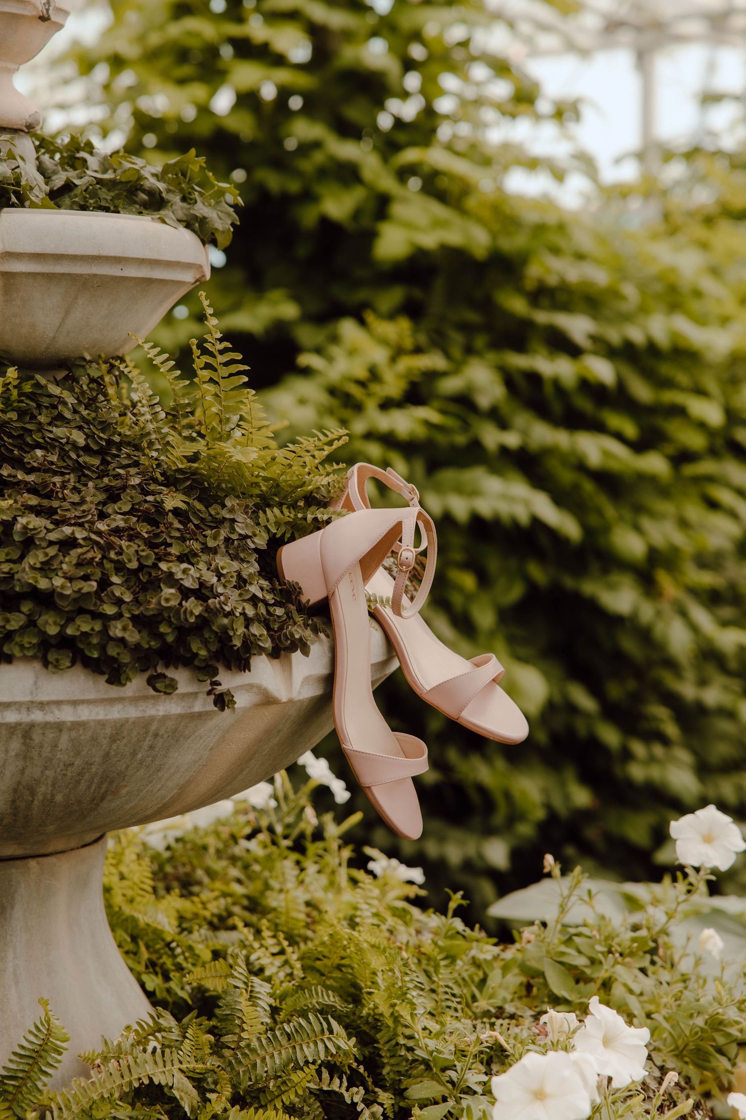 Blush pink block heel bridal sandals hanging on a stone garden fountain surrounded by lush ferns.