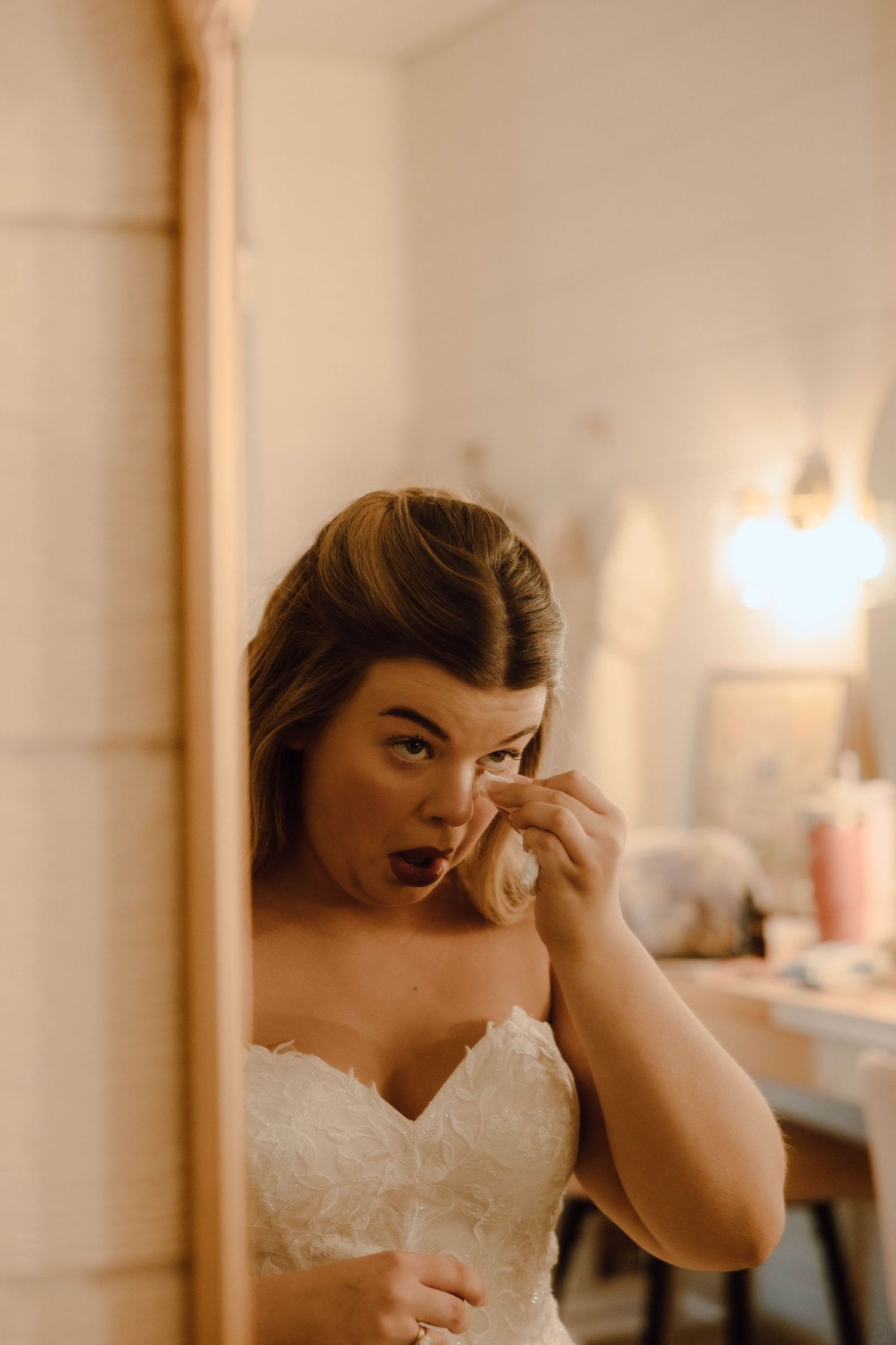 Bride in white strapless gown touching her face with surprised expression, reflected in a vanity mirror with warm lighting.