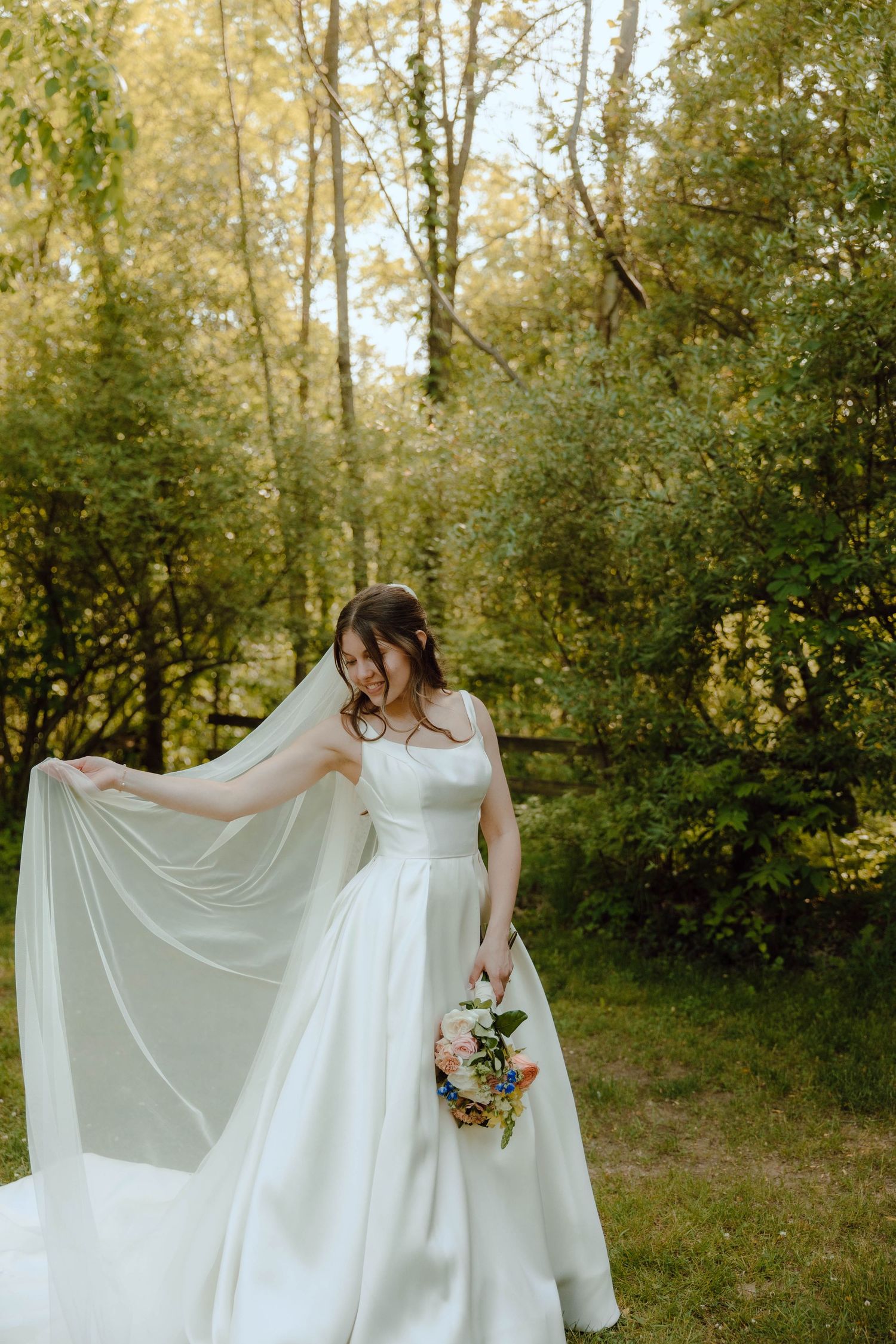 Smiling bride in sleek white gown holds colorful bouquet while her long veil flows in a lush green garden.