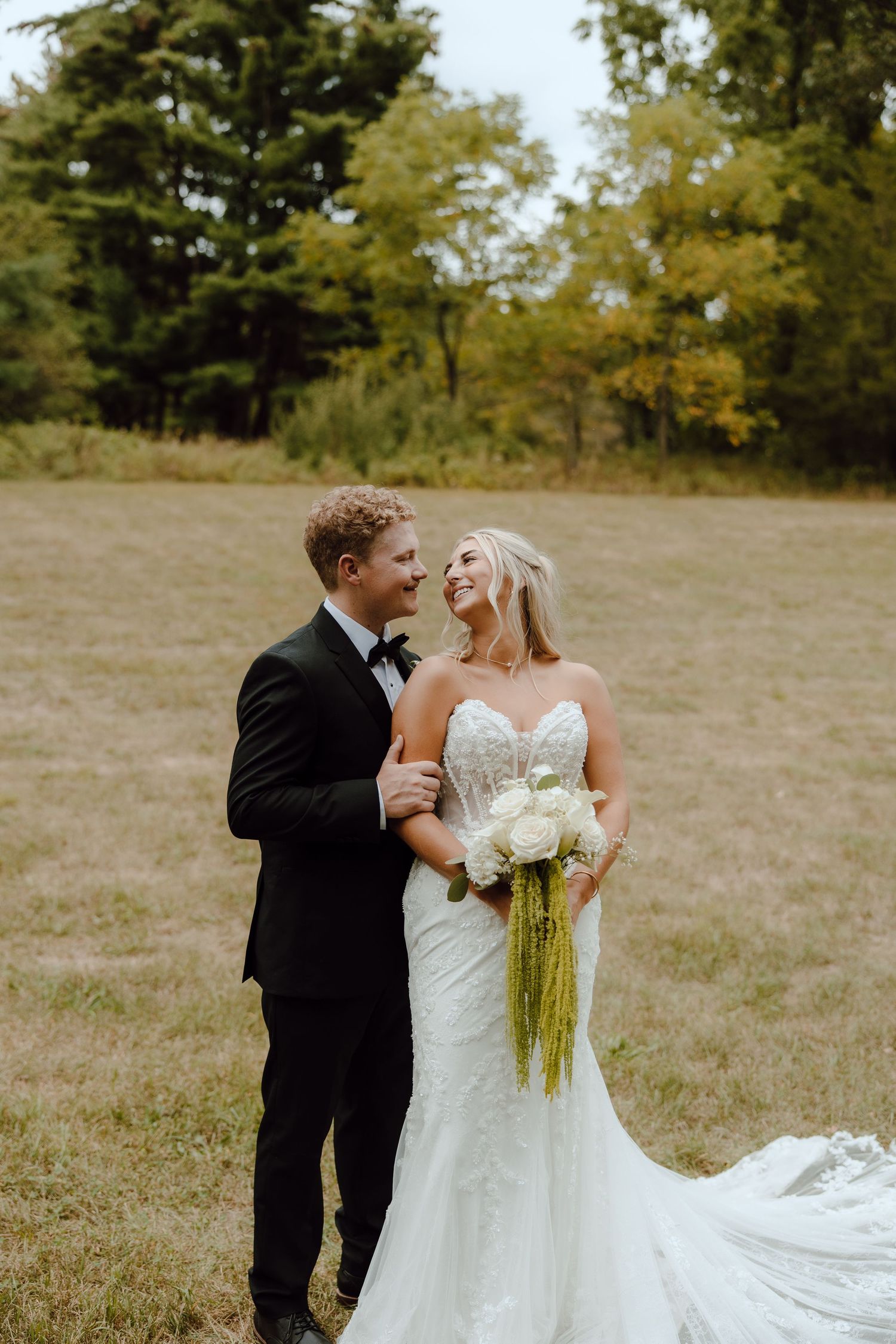 A bride and groom share a joyful moment in a field, she holds a white bouquet, he wears a black tuxedo.