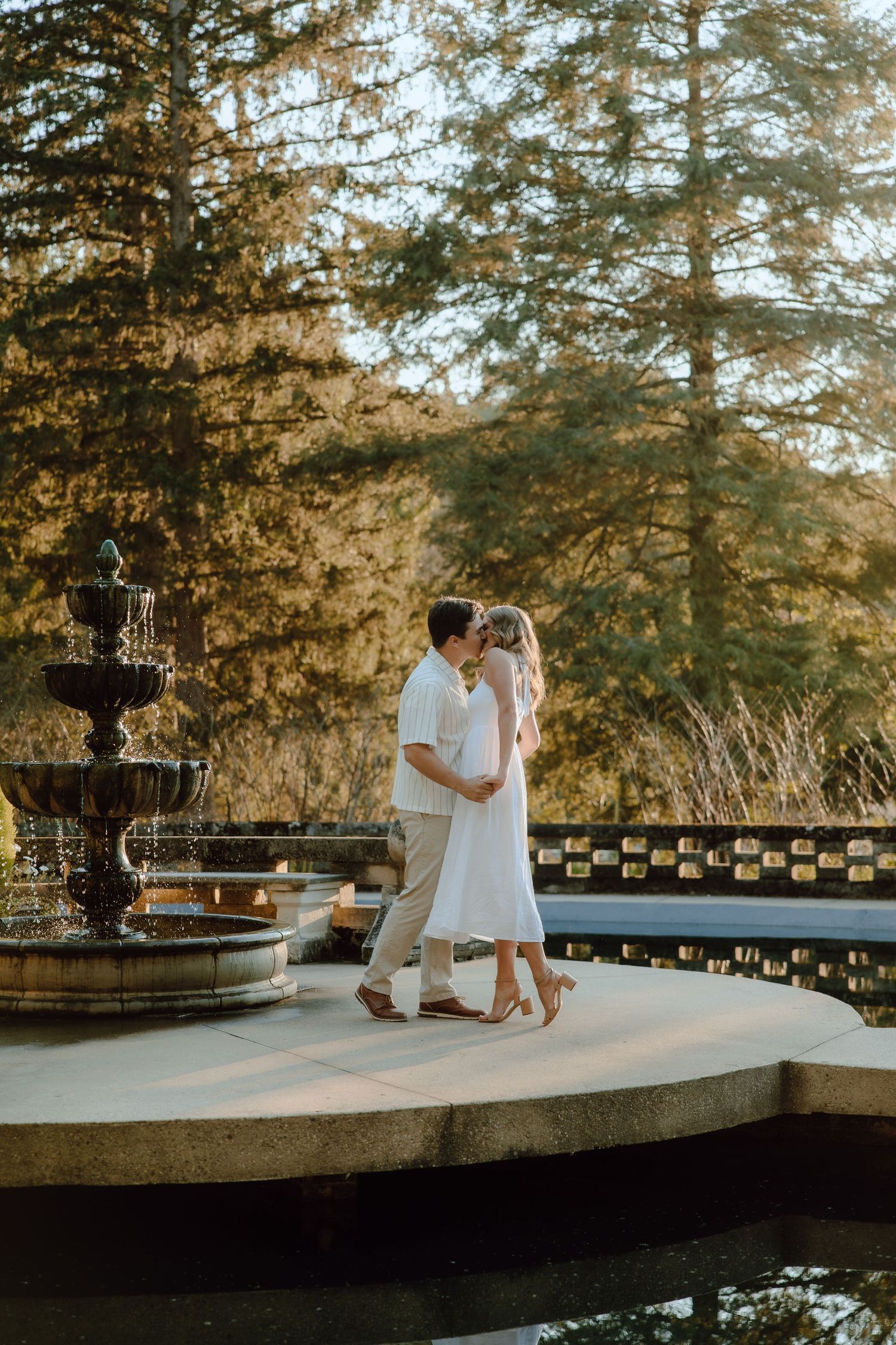 Couple sharing a romantic kiss near an ornate stone fountain surrounded by tall trees in golden light.