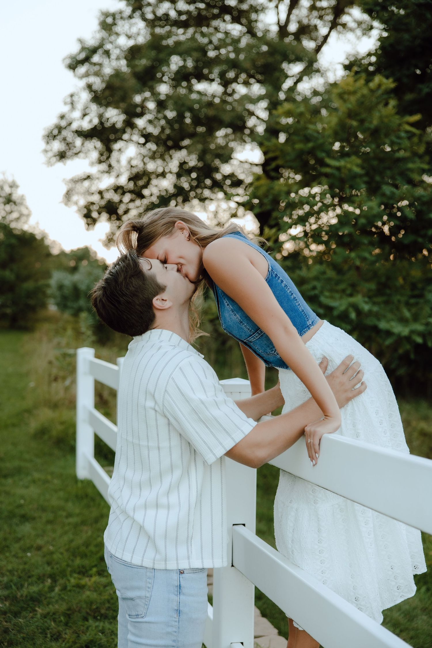 A couple shares a romantic kiss over a white fence surrounded by lush green trees in a countryside setting.