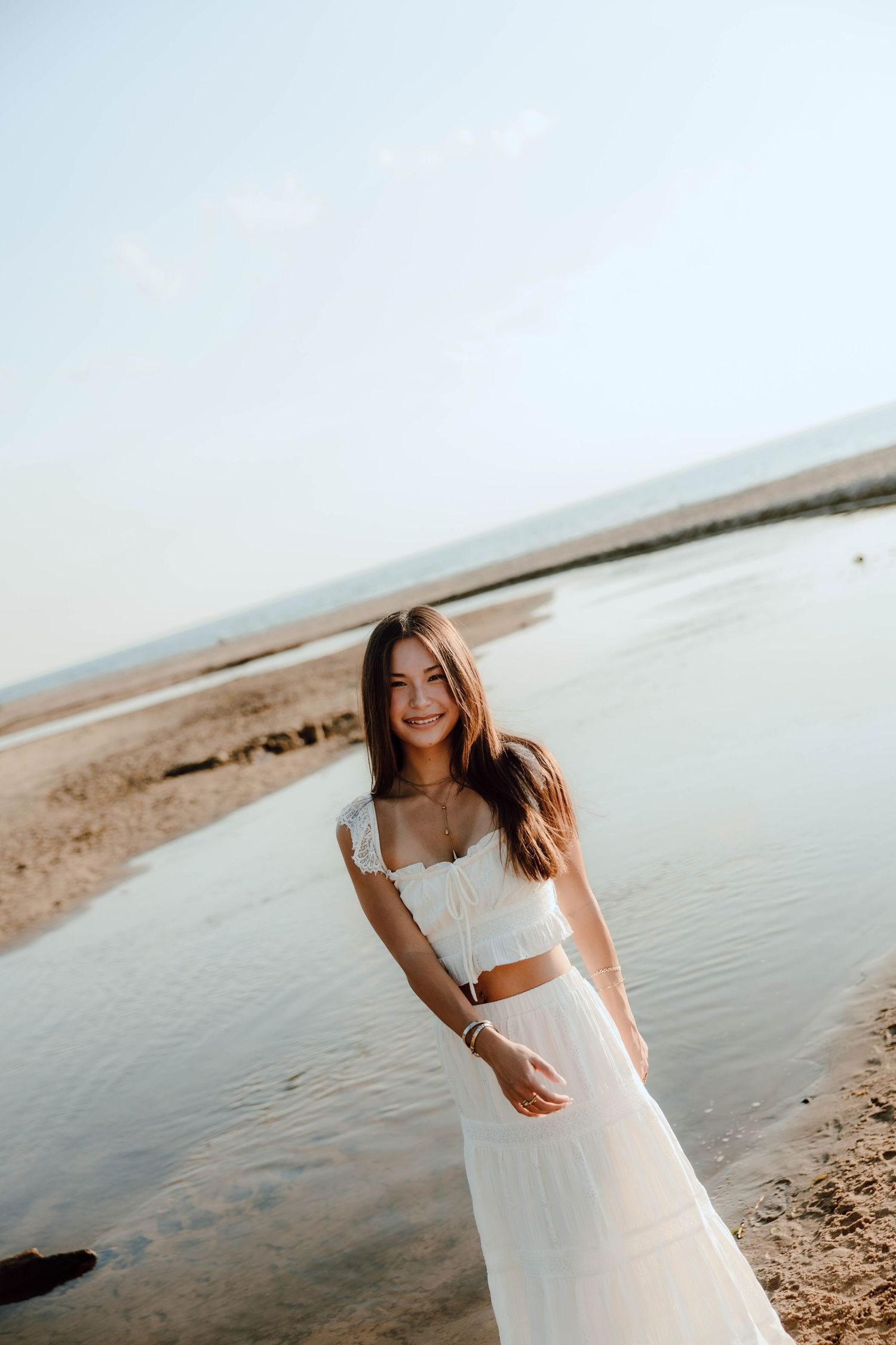 Smiling young woman in white crop top and skirt standing on sandy beach shore with calm water background.